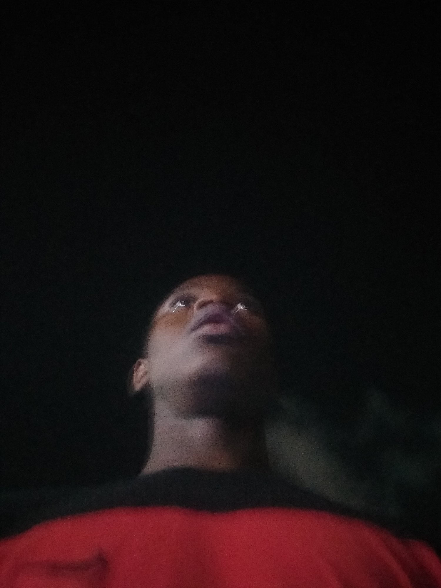 A low-angle shot depicts a person with dark skin looking upward. The subject is wearing a red shirt with a black neckline. The background is dark, obscuring any identifiable elements, but the location provided is Shangisha, Nigeria. The person's face and neck are visible, with light reflecting in their eyes. The person's attention is directed toward something out of frame above them.
