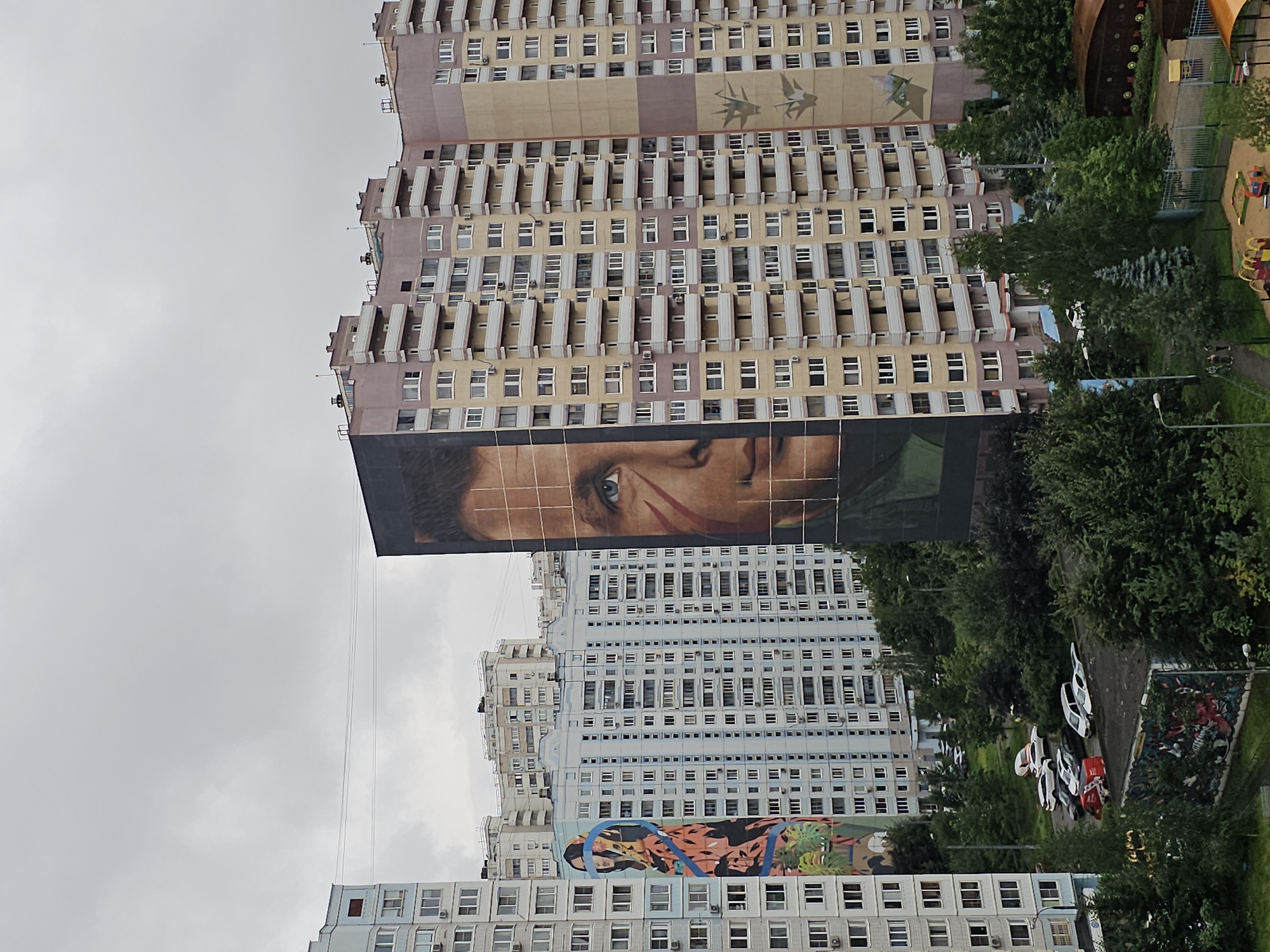 The image shows a view of residential buildings in Odintsovo, Russia, under an overcast sky. The buildings are tall, apartment blocks with many windows and balconies. One building on the right features a large mural of a man's face, with a scar across his cheek. Another building on the left has a mural depicting a woman's head and torso with colorful patterns and plants. Below the buildings, there is a street with several parked cars, and areas of greenery including trees and bushes. In the foreground, a section of a playground is visible, with sandboxes and colorful play equipment. The scene suggests a typical urban residential environment.