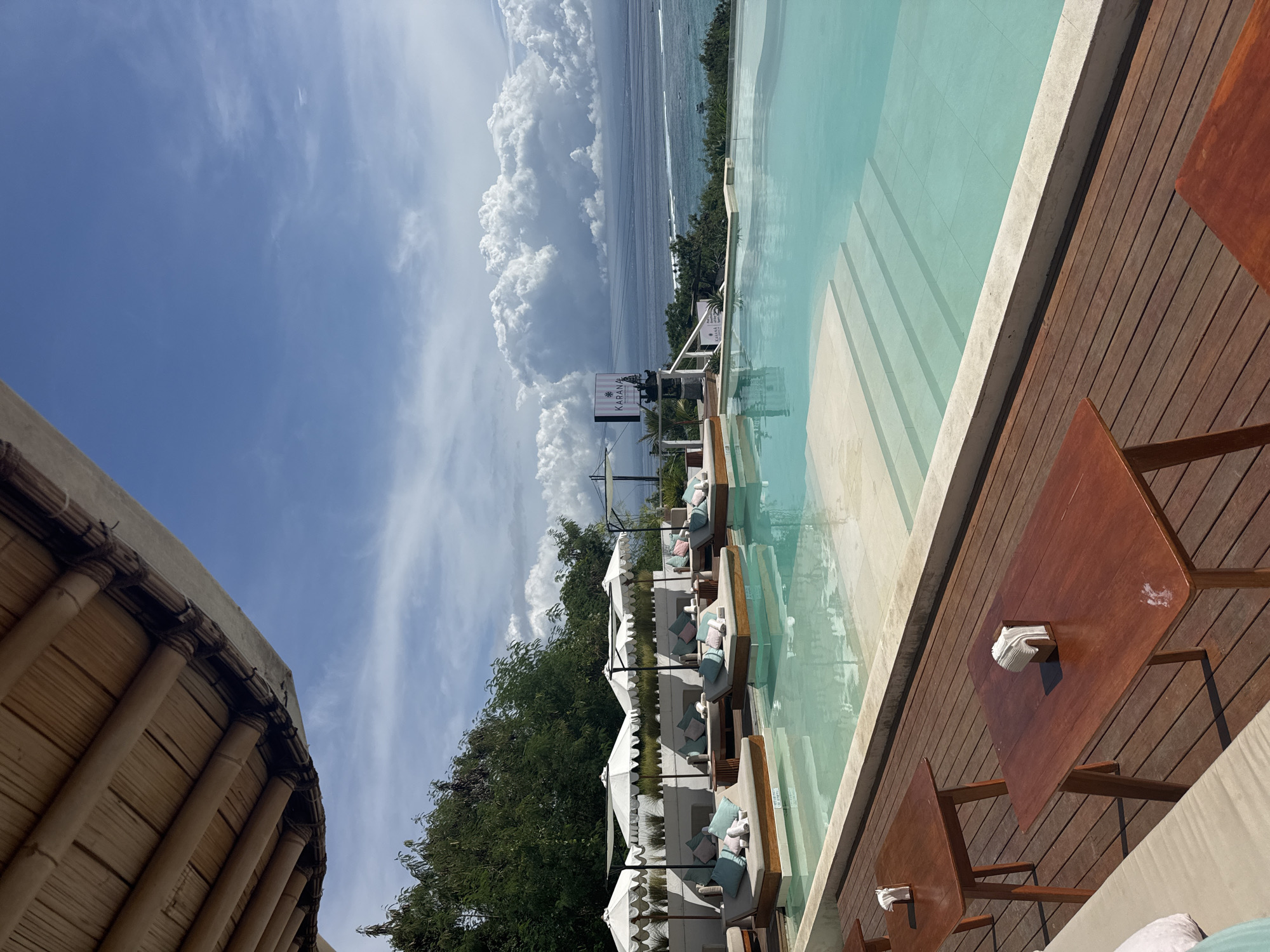 This image captures a view from a beach club in Nusa Penida, Indonesia. A thatched-roof structure is visible in the bottom left corner, suggesting an open-air setting. Adjacent to the structure is a wooden deck area with tables. Next to the deck is an infinity pool with pale-blue water. Beyond the pool, there are lounge chairs arranged in a row, each with a white sun umbrella. Lush green trees partially obscure the view towards a cloudy sky and the ocean. A sign with "KARANA" on it is visible near the lounge chairs. The cloudy sky indicates the weather conditions. The ocean is calm.