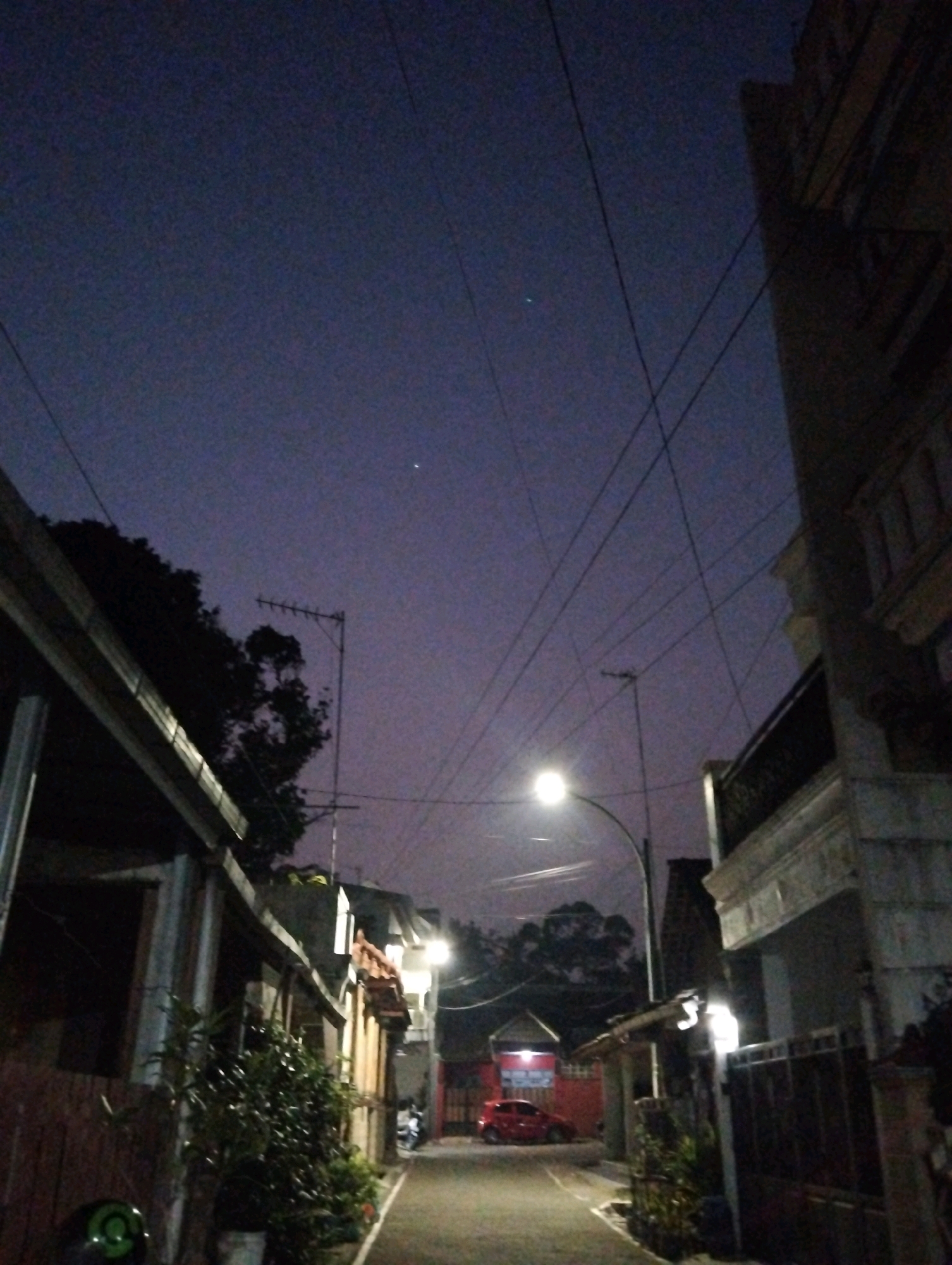 A street scene in Salatiga, Indonesia, at dusk or dawn, captures a narrow residential street lined with buildings and trees. A single streetlamp illuminates the paved road, casting a warm glow. A red car is parked on the left side of the road, and a motorcycle is visible further down the street. Power lines crisscross the sky, and a television antenna is mounted on a pole. The sky is a deep twilight blue, with a faint hint of purple. The overall impression is one of quietude and everyday life in a Southeast Asian city.