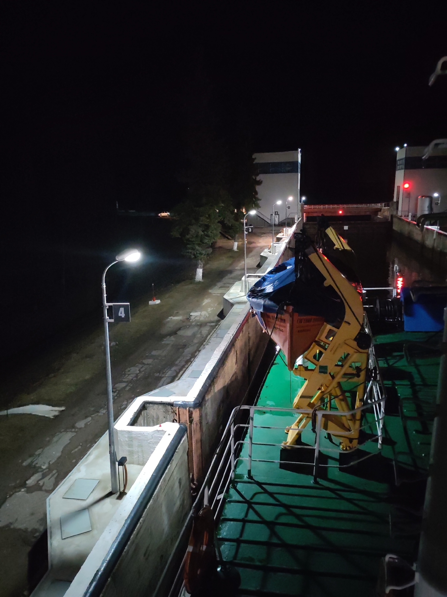 The image depicts a night scene at a canal lock in an unknown city. The view is from the deck of a vessel, showing part of the vessel's railings, deck, and a crane-like structure with a lifeboat. The lock walls are visible on both sides of the vessel, illuminated by streetlights. A service road runs alongside the canal. In the distance, there's a small structure with a red light, and a taller, white structure behind it. The night sky is dark.
