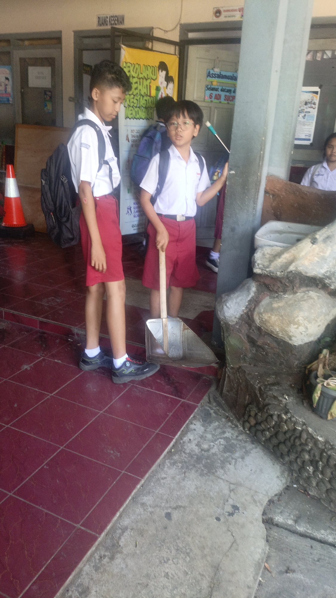 The image depicts two young male students in school uniforms standing on a tiled floor. The student on the left wears a white short-sleeved shirt, red shorts, white socks, and black shoes, and carries a black backpack. He is looking down. The student on the right, wearing similar attire, holds a dustpan with a wooden handle. He is looking forward and holding a stick with a blue end in his right hand. Behind them, a poster with Indonesian text, possibly related to school activities, is visible. The environment appears to be an outdoor area, possibly near a school building in Salatiga, Indonesia, as suggested by the text on the poster and the overall setting. The background shows a wall with doors and a decorative stone structure on the right.