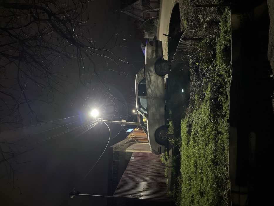 A silver pickup truck is parked on a dark street next to overgrown greenery. A street light illuminates the scene, casting a glow on bare tree branches against a dark sky. The truck's tires are visible, and a red metal structure peeks out from behind the vehicle. Power lines stretch from the street light, adding to the urban landscape. The scene is captured at night with a low angle, emphasizing the height of the light post.
