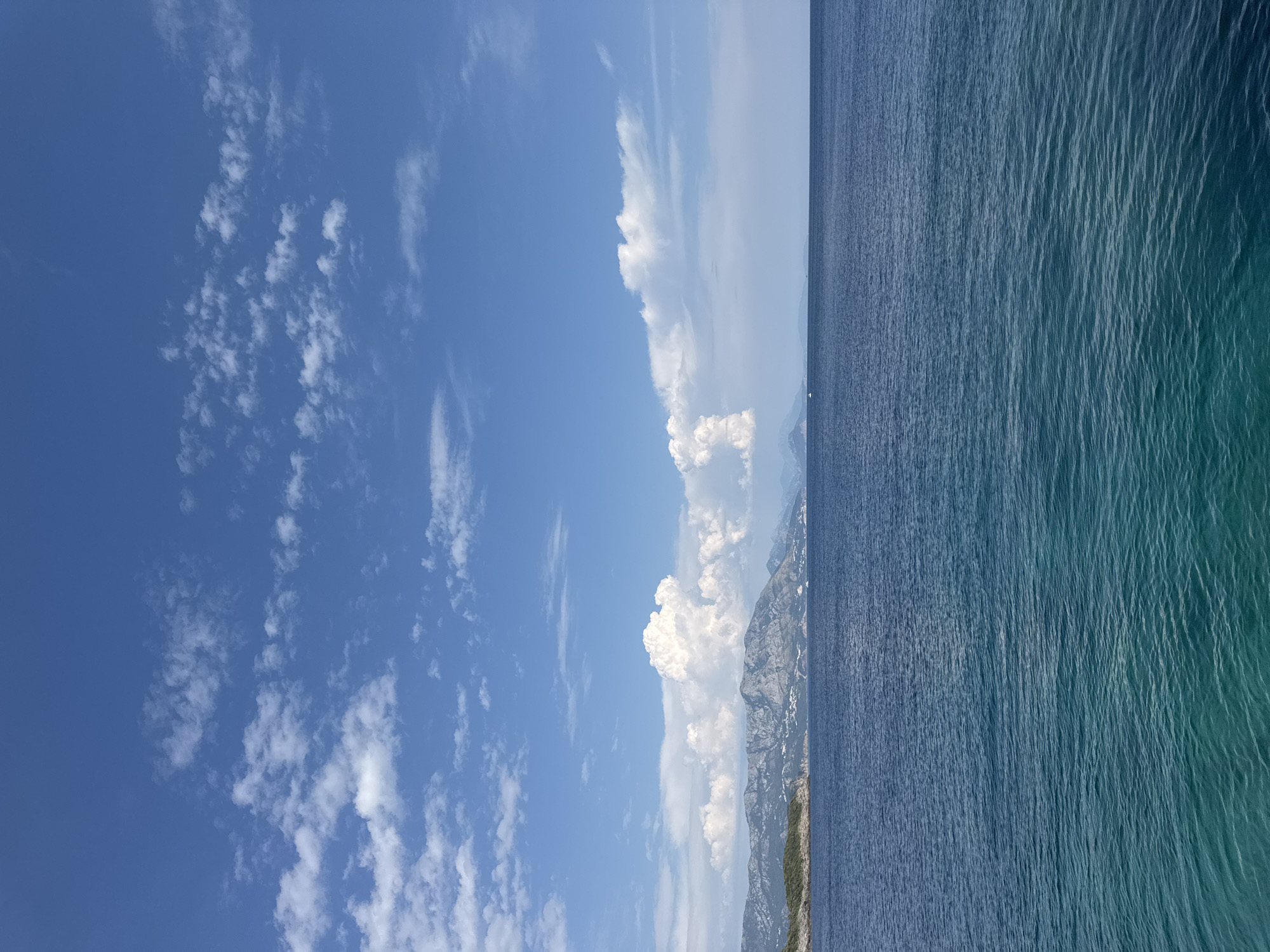 The image depicts a serene seascape with a view of the coast and mountains. The foreground is dominated by the calm, deep blue waters of the Adriatic Sea, transitioning to a lighter turquoise hue closer to the shore. Gentle ripples disturb the water's surface. In the midground, a rugged coastline is visible, dotted with sparse greenery and some scattered white structures, suggesting a settlement. Behind the coast, a range of majestic mountains rises, their peaks partially obscured by a scattering of white cumulus clouds against a bright blue sky. The clouds are dispersed, with some larger, more voluminous formations contrasting with thinner, wispy streaks. The scene is bathed in natural daylight, creating a peaceful and picturesque atmosphere. There are no people or discernible actions visible in the image. The location is identified as Dubraljevina, Montenegro, which is known for its coastal beauty and mountainous terrain.