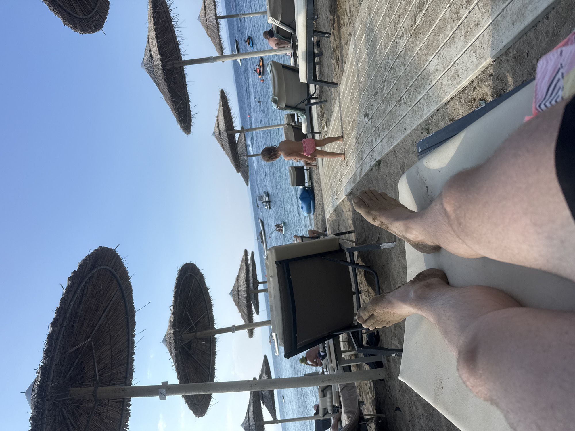 The image depicts a beach scene on a sunny day in the Sithonia Municipal Unit, Greece. The perspective is from someone lying on a beach chair, with their legs and feet, covered in sand, prominently in the foreground. Behind them, several straw umbrellas provide shade for beach chairs and lounge areas. Further back, the blue sea is visible with several boats, including a jet ski and a small boat with people, on the water. A young child in pink swim trunks stands on a wooden walkway near the water. The overall environment suggests a relaxing day at a resort or public beach.