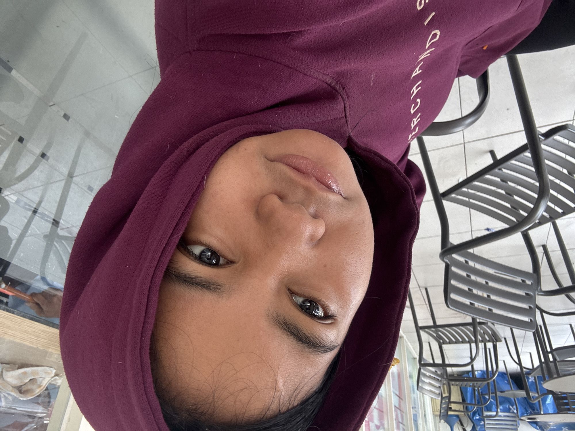 The image is a close-up, angled shot of a young woman wearing a maroon hooded sweatshirt. The woman's face is the primary focus, with her eyes looking towards the camera and her lips slightly parted. She is wearing light-colored lipstick. Her hair is dark and pulled back. The background is blurred but appears to be an indoor seating area with several dark metal chairs and tables visible. Some of these chairs are in the foreground. The floor is tiled in a light color. There are reflections on a glass surface to the right of the woman, suggesting a window or a glass partition. The text "MERCHANDISE" is visible vertically on the front of her sweatshirt. The scene is indoors, possibly a cafe or a waiting area, in North Jakarta, Indonesia.