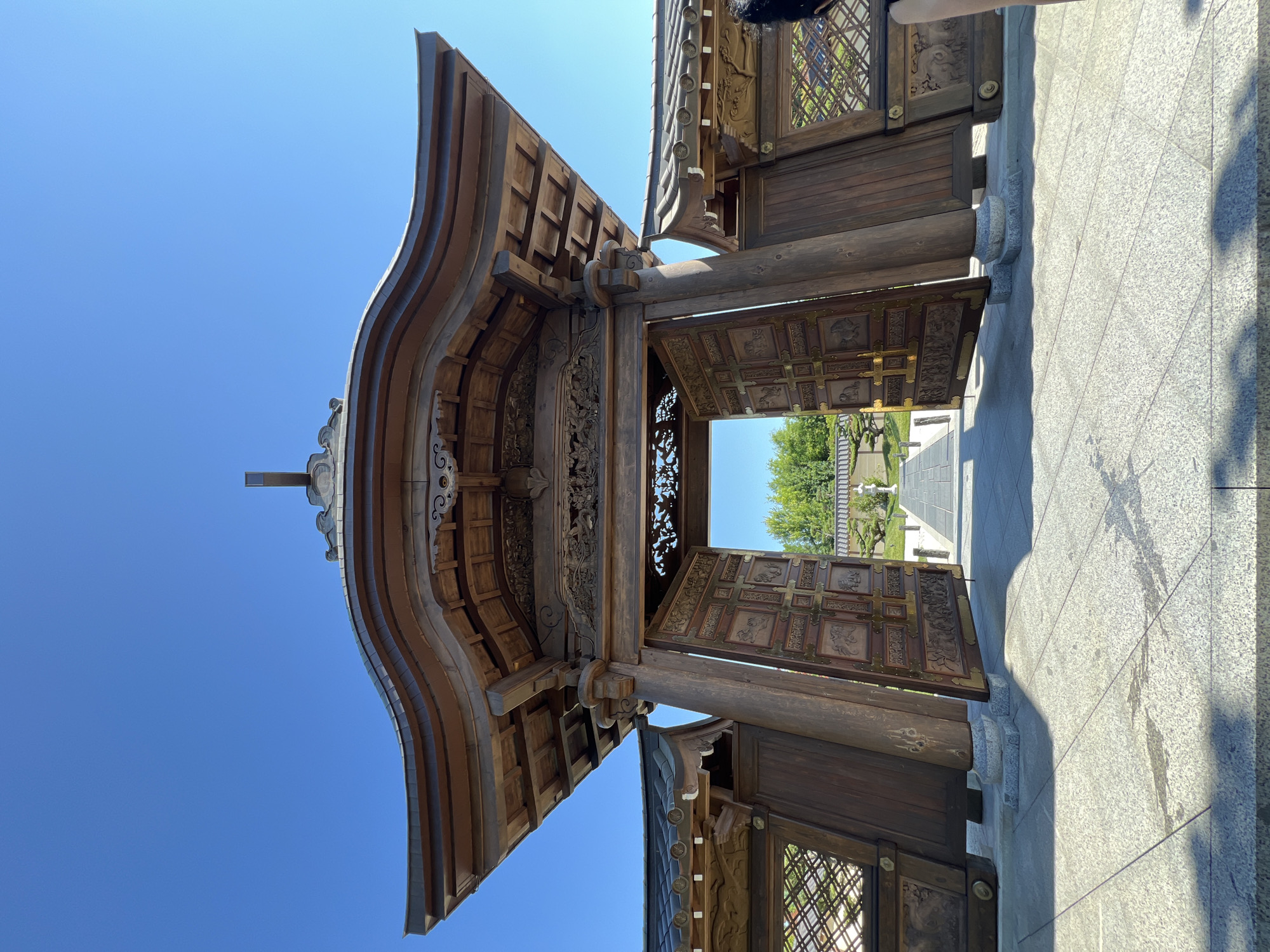 The image shows a large, ornate wooden gate in front of a temple-like structure. The gate is open, revealing a pathway leading into a garden with trees and decorative elements. The architecture of the gate and building features intricate carvings and a distinctive, multi-layered roof. The foreground consists of a paved courtyard. The sky is clear and blue. Based on the architectural style and the presence of a pagoda-like element on the gate, this appears to be a Japanese-style garden and temple. It is highly unlikely that this location is in the city of Krasnodar, Russia, as the architectural style is not indigenous to that region.