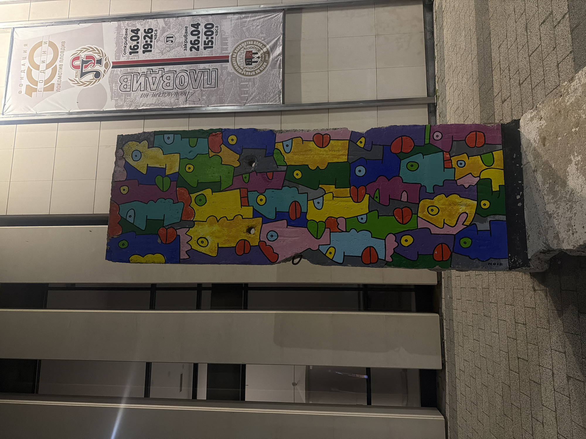 The image shows a vibrant, abstract mural displayed outdoors beneath a large commemorative banner. The mural features a dense, interlocking pattern of stylized, colorful figures resembling fish or fantastical creatures, rendered in bright shades of yellow, blue, green, purple, pink, and red. Each figure has distinct eyes, and one notably has a circular hole. The artist's signature, "NOIR," is visible in the bottom right corner of the mural.
Above the artwork, a banner advertises an event celebrating "100 ГОДИНИ" (100 Years) of "ЛОКОМОТИВ ПЛОВДИВ" (Lokomotiv Plovdiv), founded in "1926." The banner includes the club's logo with "ЛП" and mentions "Любимецът на ПЛОВДИВ" (The darling of Plovdiv). Event details specify an "откриване" (opening) on 16.04 at 19:26 and a "закриване" (closing) on 26.04 at 15:00. Another logo for a "КЛУБ НА ПРИВЪРЖЕНИЦИ" (Club of Supporters) is also visible.
The setting appears to be an urban exterior, possibly part of a building facade or a public underpass. Below the mural, horizontal architectural elements and windows are visible, reflecting artificial light, suggesting the time of day is evening or night. To the right, a textured wall made of light-colored tiles or stone completes the scene, which combines public art with a community-focused commemorative event.