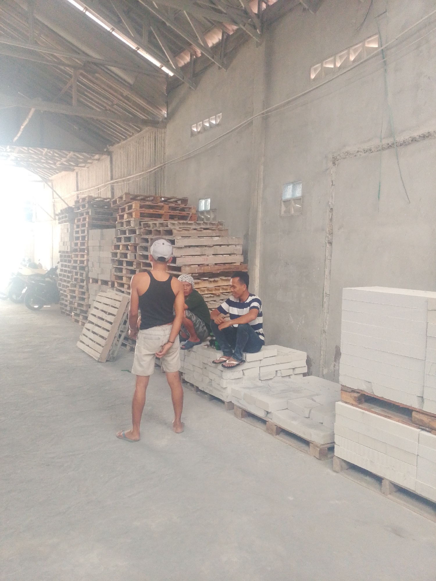 The image depicts an indoor scene of a warehouse in Banjarmasin, Indonesia, likely a distribution or storage facility for building materials. In the foreground, a person wearing a black tank top, light-colored shorts, and a grey cap stands with their back to the camera, facing a stack of wooden pallets. To the right, two other individuals are seated on stacks of white blocks. One person wears a horizontally striped t-shirt and dark pants, while the other is partially obscured, wearing a green shirt and camouflage pants. The warehouse interior is characterized by a concrete floor, tall concrete walls with a few small windows or ventilation blocks, and a wooden beam ceiling. Stacks of wooden pallets and white building blocks are prominent throughout the space. In the background, several motorcycles are visible, suggesting a means of transportation or delivery. The overall atmosphere is one of industrial activity and storage.