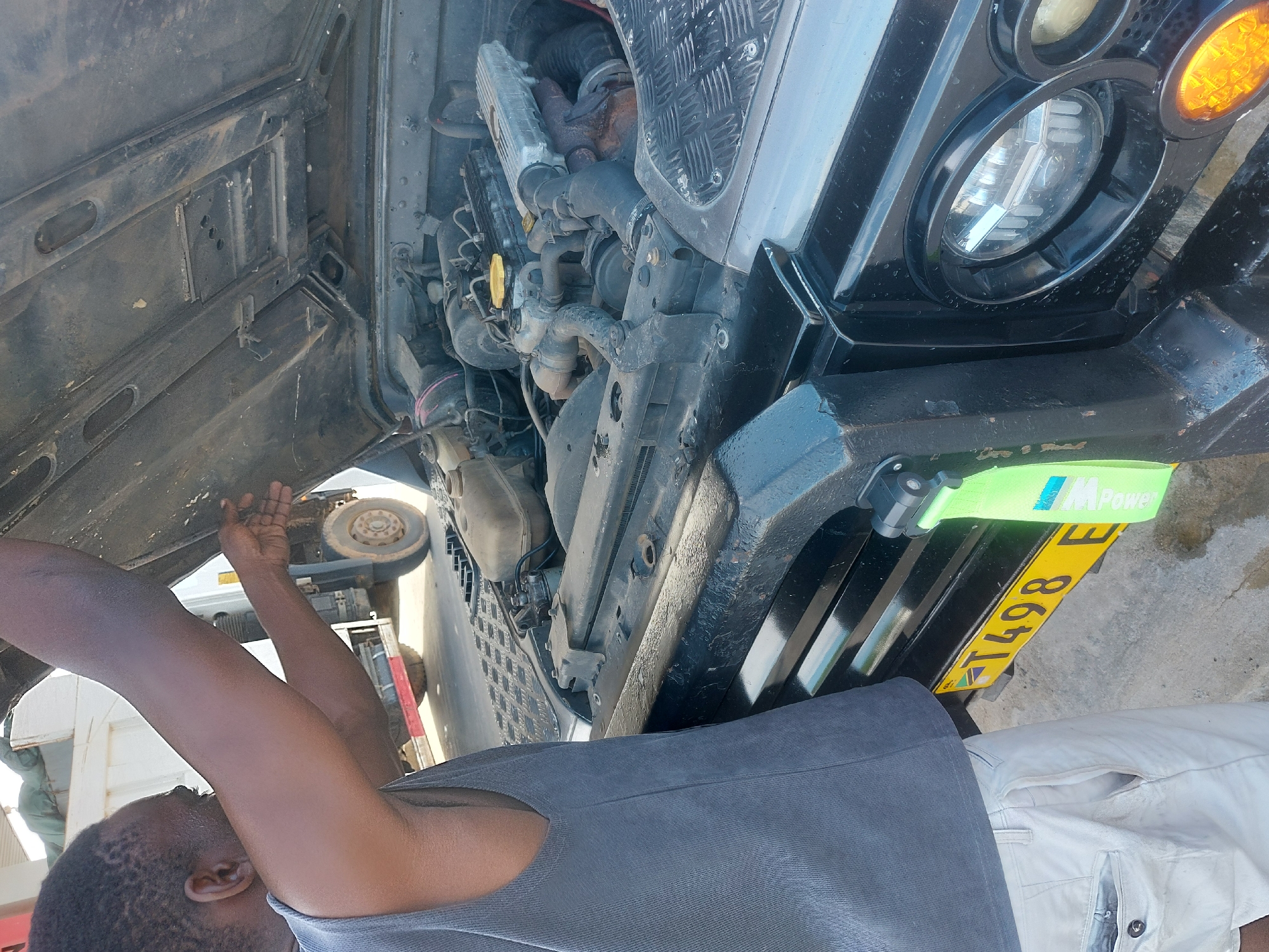 The image shows a man working on the engine of a car. The car appears to be a Land Rover Defender, judging by the distinct front-end design and engine layout. The hood is open, revealing the engine compartment and its components. The man is reaching into the engine bay, presumably to perform some maintenance or repair work. The vehicle has a Tanzanian license plate reading "T498 E". Considering the license plate and absence of other visual context, it is not possible to confirm the location within the city of Dar es-Salaam, Tanzania, or provide a definitive forensic location.