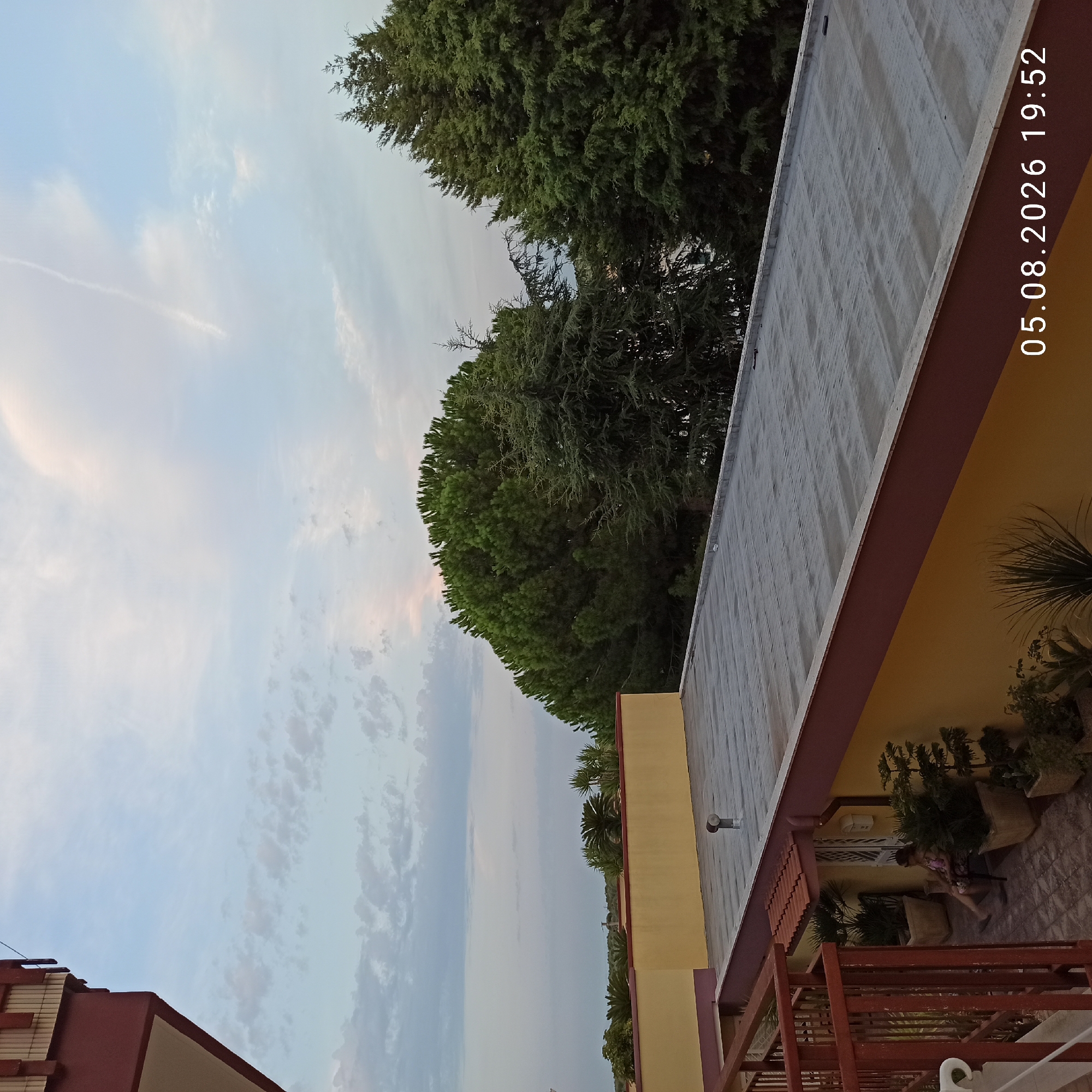 The image shows a rooftop view in the city of Bitonto, Italy, with a timestamp of "05.08.2026 19:52". In the foreground, a woman is seated on a chair on a patio area, surrounded by potted plants and palm trees. A red railing is visible to the left, suggesting a balcony or terrace. The building features yellow walls and a dark red trim. Beyond the building, large evergreen trees dominate the landscape, with a pale blue sky with scattered clouds overhead. The overall scene depicts a quiet evening setting, possibly residential, with a focus on outdoor space and natural elements.