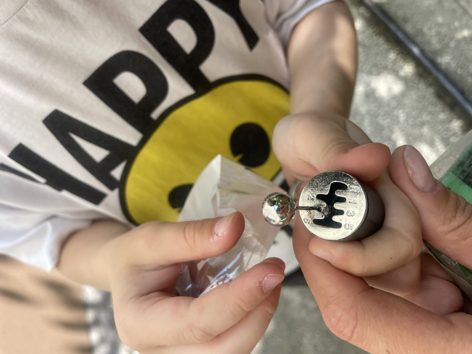 In a close-up outdoor shot, a person's hands are holding a miniature gear shift knob. The knob is silver and has the typical manual transmission shift pattern engraved on it, with numbers 1, 3, and 5 visible, along with "R" for reverse. A small silver ball sits atop the gear shift lever. The background shows a child wearing a white t-shirt with a large yellow smiley face and black lettering that reads "HAPPY." The ground appears to be a textured, light-colored surface. The scene is set in Sochi, Russia, though no specific landmarks are identifiable. The overall impression is a candid moment of play or demonstration of a novelty item.