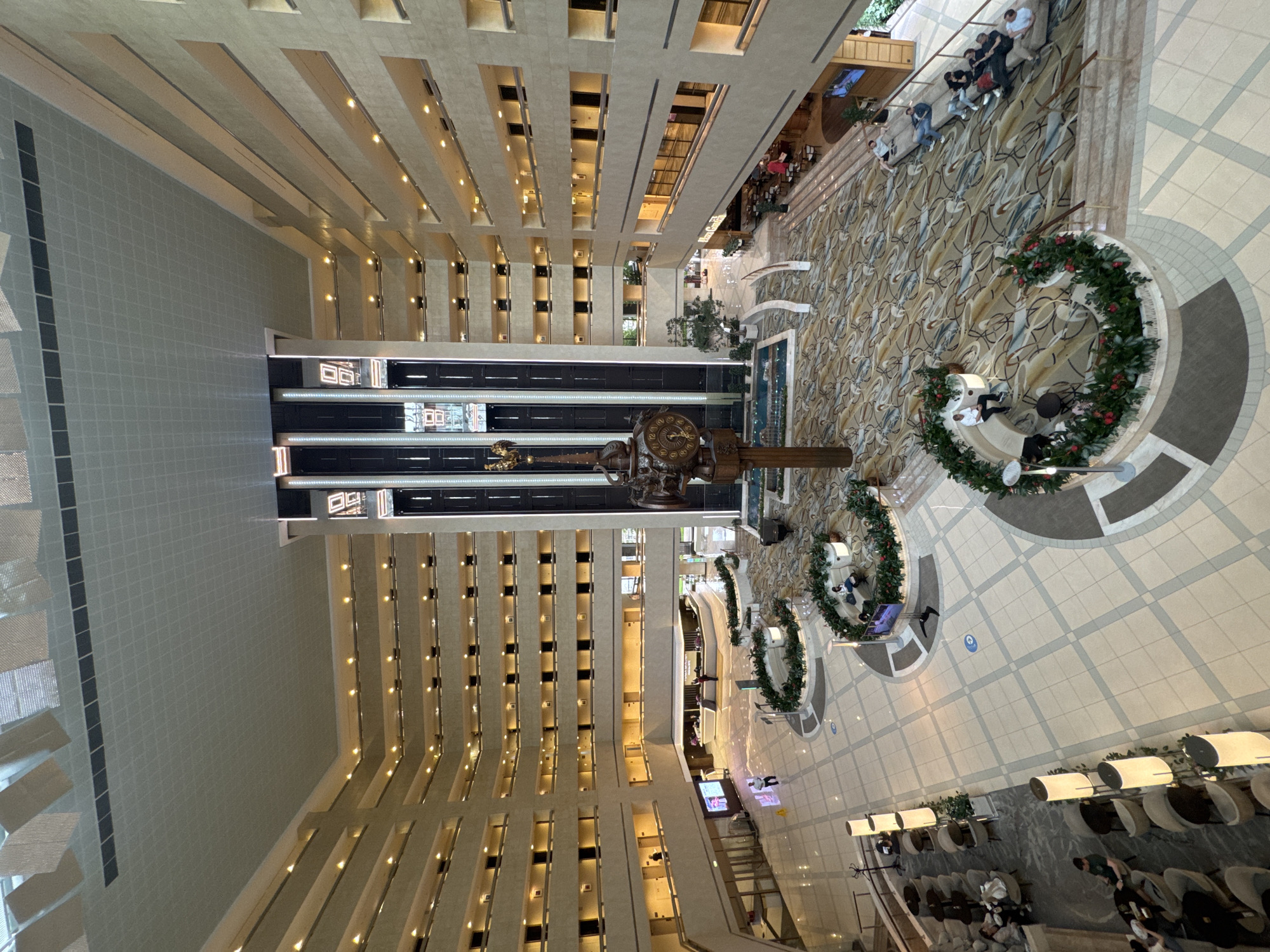 An interior overhead shot shows the multistoried lobby of a large hotel with rows of guest room doors visible on many floors, on either side of a central atrium. Near the center of the atrium is a large, ornate clock sculpture atop a water fountain. Seating areas with decorative wreaths are placed on either side of the fountain. Further away, near the building's edges, are additional seating areas and a restaurant. People are present in the various public areas, including the restaurant and seating areas. The color scheme throughout is mostly neutral with accents of black, gold, and green.