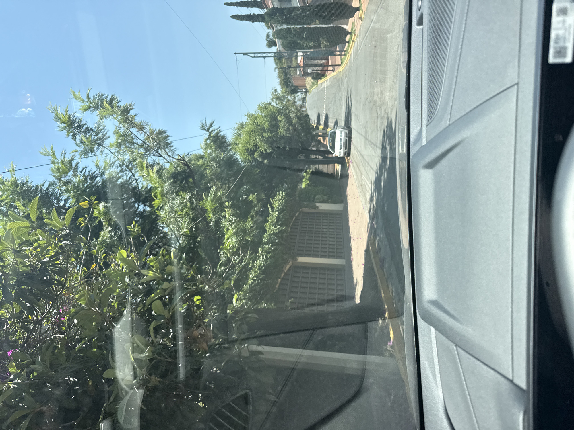 The image, presented in a landscape orientation but likely captured vertically, displays a residential street scene in San Bernardino Tlaxcalancingo, Mexico, viewed from inside a vehicle.
The right side of the frame is occupied by the dark gray interior of the vehicle, showing a textured door panel and part of the dashboard, indicating the photo was taken through a windshield or side window.
Outside the vehicle, a paved road gently curves and ascends into the distance. Yellow lines mark the curb on the right side of the road. A white sedan is parked on the right side of the street, facing away from the viewer.
Lush green vegetation borders both sides of the street. On the left, dense trees and bushes are prominent, some with visible purple flowers. On the right, tall, slender, conical trees (resembling cypress) line the street, with glimpses of buildings and a metal gate behind them. Overhead, power lines and a utility pole are visible against a clear, bright blue sky, suggesting a sunny day.
No people are visible in the scene. The overall impression is a quiet, tree-lined residential area during daytime.