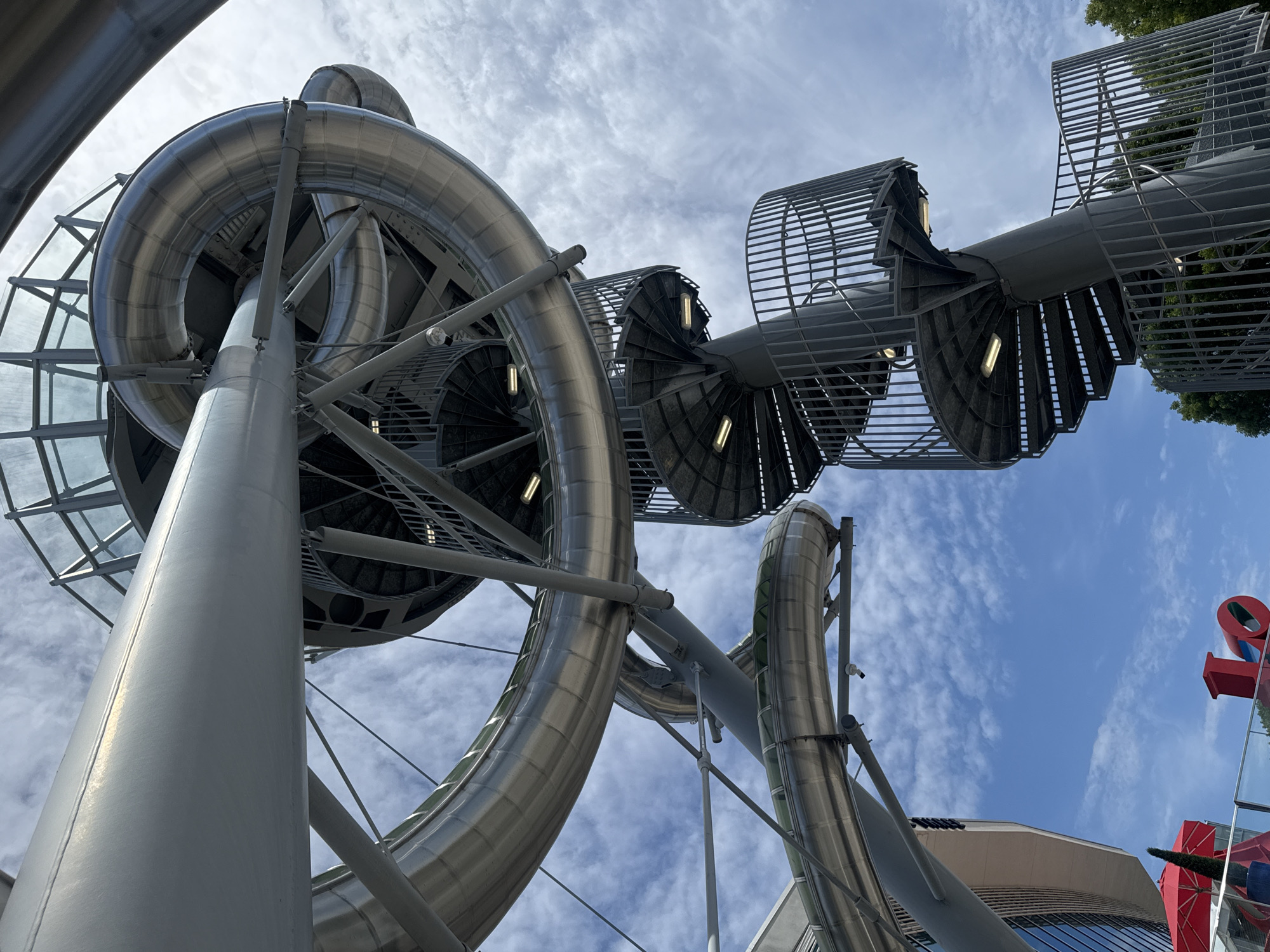 This image captures a low-angle view of a large, metallic, spiral slide structure set against a bright blue sky with scattered clouds. The slide is made of gleaming silver material, with transparent sections revealing glimpses of the interior. Multiple circular staircases with metal railings spiral upwards, connecting to different levels of the slide. The stairs are illuminated by modern, rectangular lights.
In the background, to the left, a modern building with glass windows is visible. Below the slide structure, a red umbrella and a red sculpture that appears to spell "LOVE" are partially visible.
This structure is located at Aventura Mall in Aventura, Florida, United States of America, and is part of the mall's architectural design and recreational amenities. There are no people visible in the image.