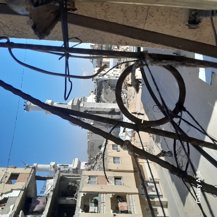 The image displays a scene of significant urban destruction in Khan Yunis, Palestinian Territory, captured from an elevated perspective through rusted metal bars and wires. The sky is clear and bright blue.
On the left, a multi-story building exhibits severe structural damage, with multiple floors partially collapsed and debris visible within its exposed sections. A single individual is discernible on a balcony of this damaged building. The street below, composed of asphalt interspersed with exposed earth and rubble, shows signs of disarray. Three individuals dressed in dark clothing are observed walking away from the viewer on the left side of the street. On the right side of the street, a person is riding a horse or donkey, moving towards the right.
In the mid-ground and background, a large white mosque with two distinct, largely intact minarets is prominent. However, the mosque's central structure and numerous adjacent buildings are heavily damaged or reduced to rubble, contributing to a landscape dominated by debris and collapsed structures.