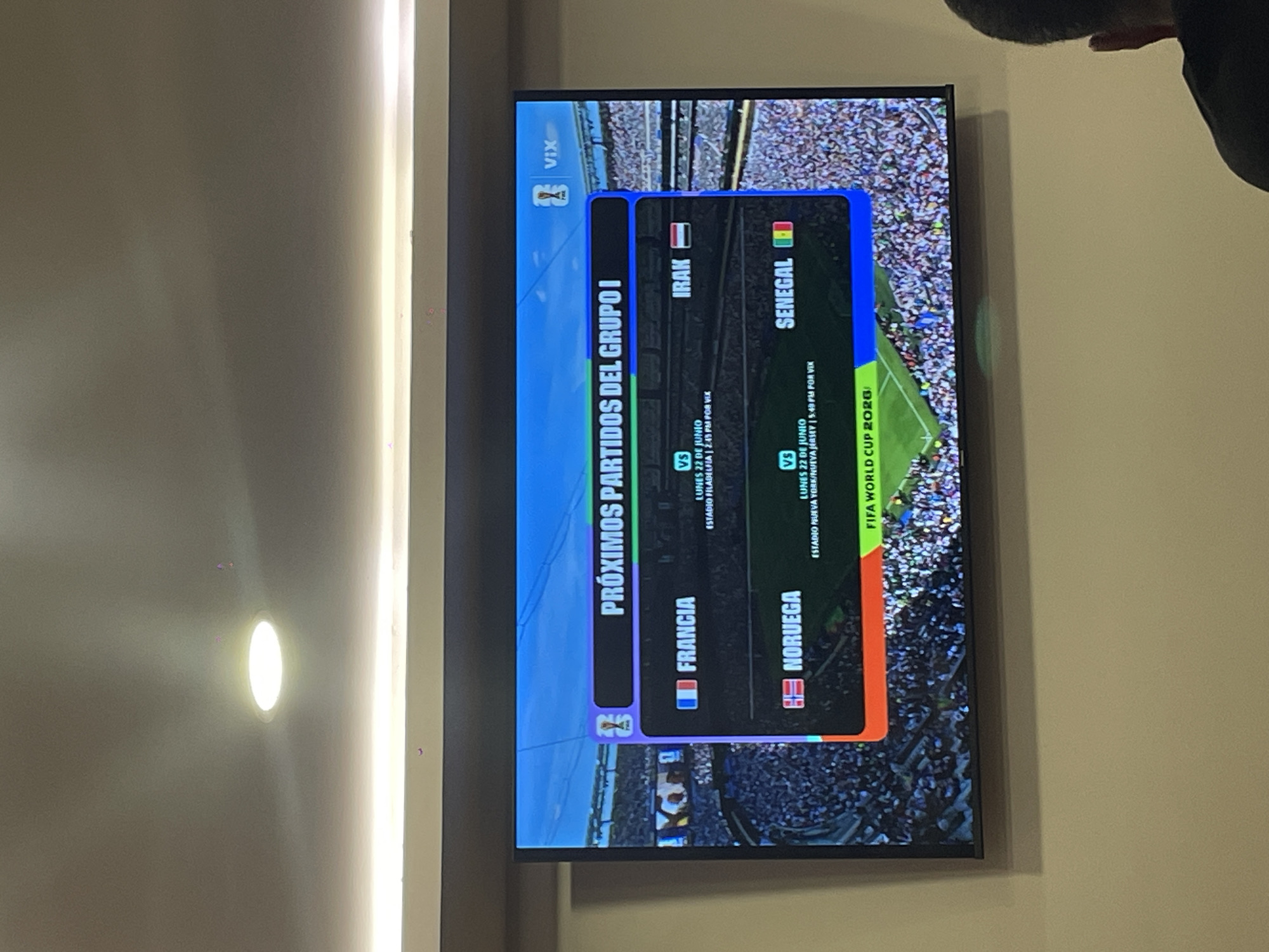 The image shows a television screen displaying upcoming matches from "Grupo I" of the FIFA World Cup 2026. The screen exhibits the VIX logo and lists two matches: "Francia vs Irak" and "Noruega vs Senegal". Both matches are scheduled for "Lunes 22 de Junio" (Monday, June 22nd). Details about the stadium and broadcast time are also visible, with one match listed as being at "Estadio Filadelfia | 2:45 PM for VIX" and the other at "Estadio Nueva York | 5:49 PM for VIX". The television is mounted on a wall, with a ceiling light visible above it. In the foreground, a person wearing a black shirt is seated and partially visible from behind, facing the television. The scene appears to be indoors. Based on a logo on the screen reading "VIX", it is highly probable that this is being viewed in Mexico, given that VIX is a major streaming service in that country. While the specific location within Cuautitlán Izcalli, Mexico cannot be definitively determined from the image alone, the presence of the VIX logo strongly suggests a viewing location within Mexico.