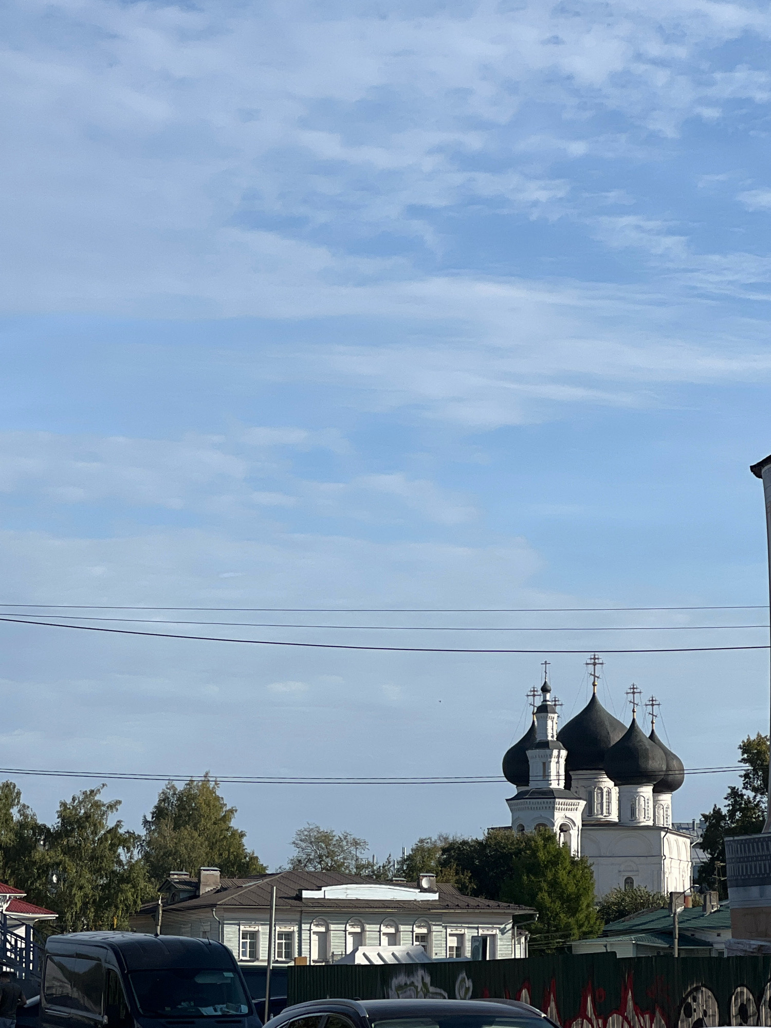 The image shows a white church with black domes and crosses, set against a blue sky with scattered clouds. The church is a prominent structure, with several smaller buildings and trees visible around its base. In the foreground, there are power lines stretched across the frame. There are also several vehicles, including a black van, and a fence with graffiti visible. The overall scene depicts an urban environment with a mix of architectural styles and natural elements.
