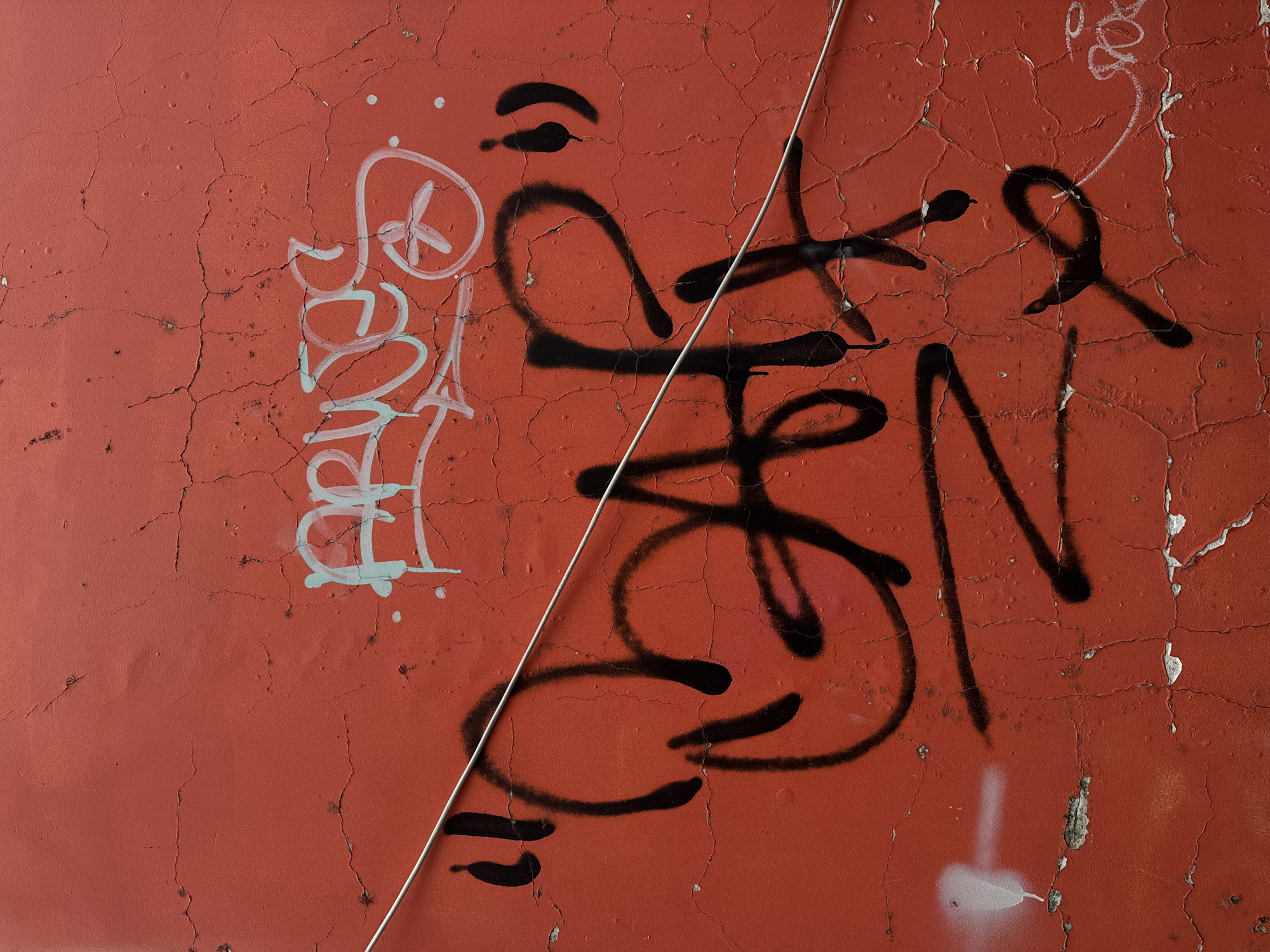 The image is a close-up shot of graffiti on a cracked, orange-red wall. A thin silver wire diagonally bisects the frame from the upper left to the lower right. In the upper right portion of the image, faint light blue graffiti with white outlines is visible, forming abstract shapes and possibly letters. The primary focus is on bold black graffiti in the center and lower portion of the frame, appearing to spell out "BAR" with quotation marks at the beginning and end. Below this, other black graffiti elements are present, including what looks like a stylized "N" and other abstract marks. A spray of white paint is on the left side of the image near the bottom. The wall's texture is rough and shows significant cracking, suggesting age or exposure to the elements. The overall impression is of urban decay and street art. The provided context places this image in Da Lat, Vietnam.