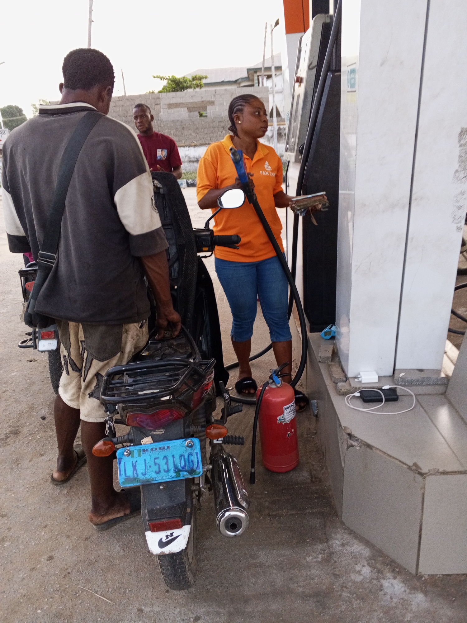 The image depicts a gas station scene with three individuals and a motorcycle. An attendant in an orange shirt is operating a gas pump. To her left, a man in a dark gray and beige shirt stands next to a motorcycle, holding what appears to be a metal carrier. Another man in a maroon shirt stands further back, looking towards the attendant. A red fire extinguisher sits near the base of the gas pump. A license plate is visible on the motorcycle, displaying "LKJ-531OG." A power adapter and a power bank connected by a charging cable sit on a ledge near the gas pump. The specific city or location is not evident in the image.