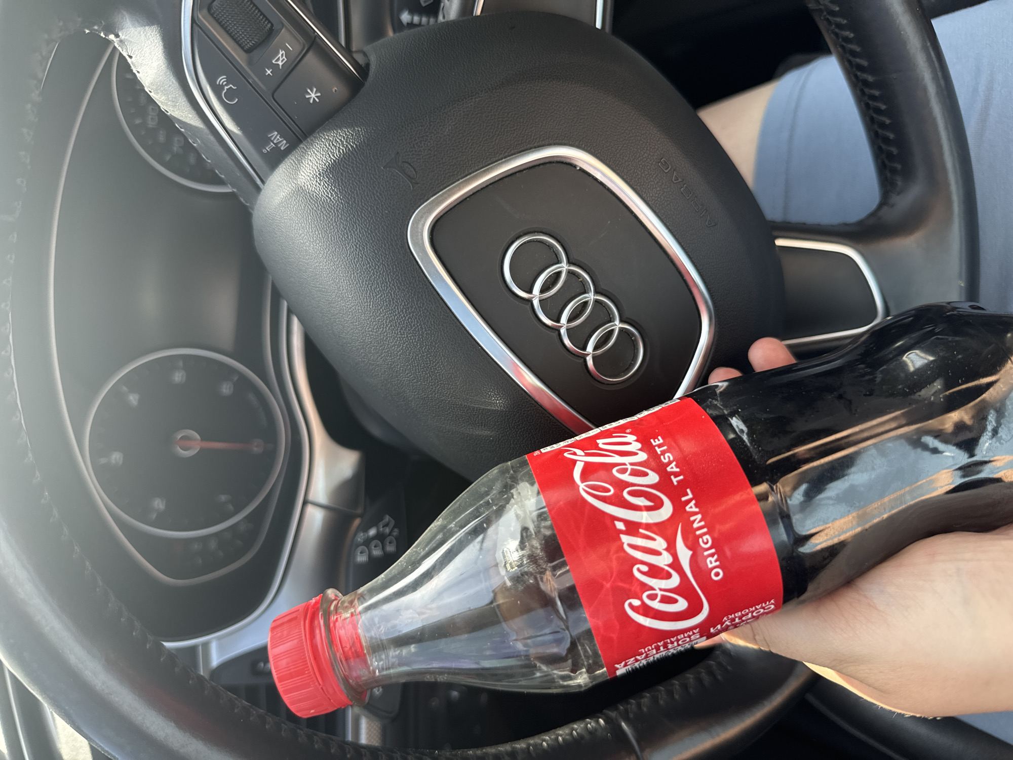 The media file shows a close-up, overhead view of a person holding a bottle of Coca-Cola in a car. The steering wheel of an Audi is prominently featured, with the brand's four rings emblem visible in the center. The dashboard gauges and steering wheel controls are also visible. The person's hand, with manicured nails, is holding the plastic bottle, which has a red cap and a red label with the Coca-Cola logo and "ORIGINAL TASTE" text. The background shows a person's legs in light blue shorts and part of the car's interior. The lighting suggests it is daytime, possibly sunny, given the bright highlights. The scene appears to be a casual moment, with the person likely taking a break or a sip of their drink while in the driver's seat. The location context is Sarny, Ukraine.