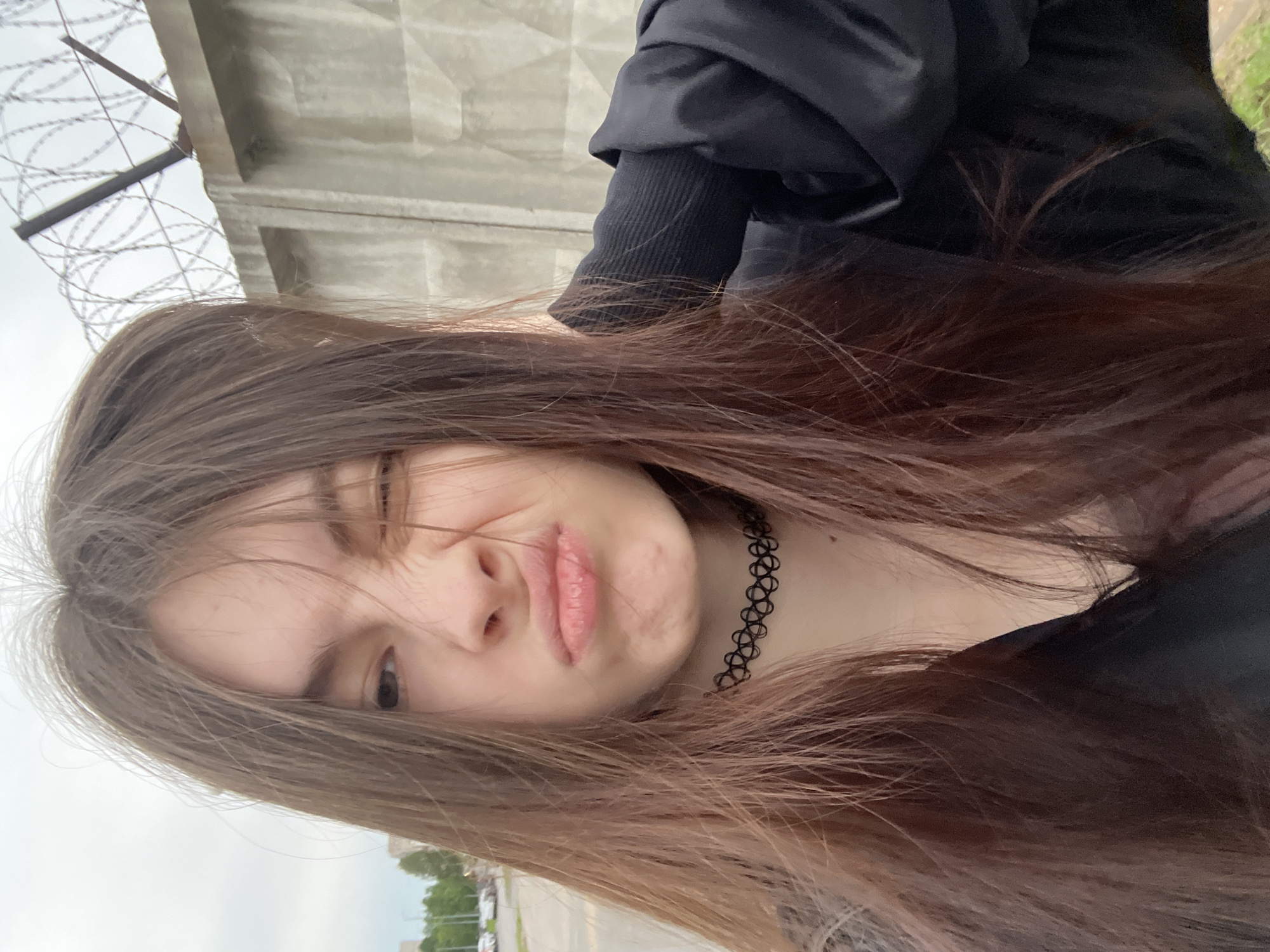 A young, fair-skinned individual with long brown hair wearing a black choker and black clothing is positioned in front of a concrete wall topped with barbed wire. The subject is making a face, squinting one eye and pursing their lips. Outside the frame, partial elements of an urban environment are visible, including a street and trees in the background. The image suggests an outdoor location, likely in an urban area with security measures (the barbed wire). There is no data in the provided content indicating Saint Petersburg, Russia.