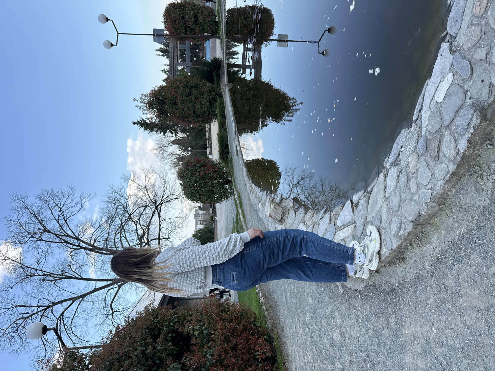 A woman is standing next to a pond in a park. The pond reflects the trees and buildings behind the woman. The park is located in Stara Zagora, Bulgaria. The woman is wearing a white sweater and blue jeans and white shoes. She is standing on a stone walkway next to the pond. The pond is lined with stones. There are trees and buildings in the background. The sky is blue with some clouds. A lamp post is visible in the background.