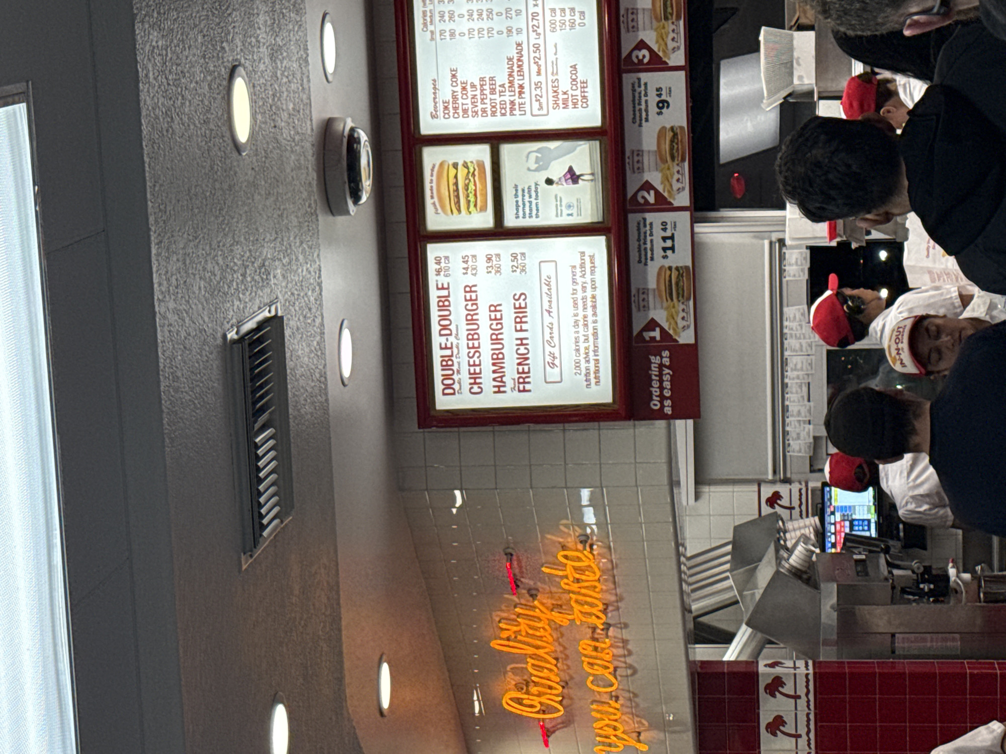 The image shows an indoor scene at an In-N-Out Burger location, likely in West Hollywood, California, as indicated by the In-N-Out brand. The focus is on the menu board displaying prices for items like the Double-Double, Cheeseburger, Hamburger, and French Fries, along with beverage options and combo meal deals. Several customers are visible, some wearing In-N-Out uniforms, suggesting they are employees. The environment includes tiled walls, lighting fixtures, and the restaurant's "Quality you can taste" neon sign. The ordering process is in progress.