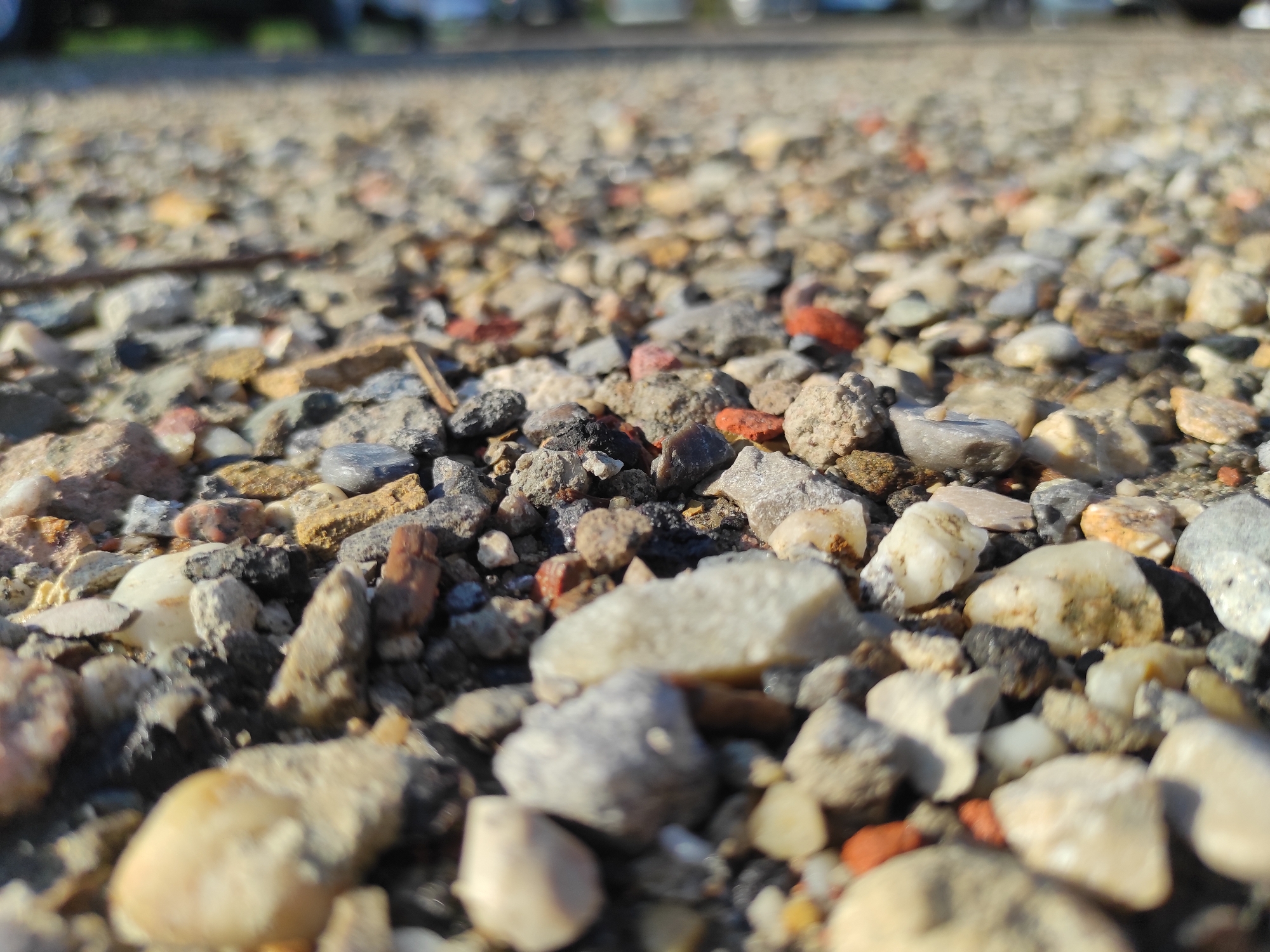 The image is a close-up, low-angle shot of a gravelly surface, likely a road or pathway, in Chaponost, France. The foreground is dominated by an assortment of small to medium-sized stones and pebbles in various shades of grey, white, brown, and reddish-orange. Some of these stones are angular, while others are smooth and rounded. Scattered among the stones are fragments of what appear to be broken bricks or concrete, particularly noticeable in the reddish pieces. The surface is uneven, with shadows cast by the stones indicating a light source from the right. In the blurred background, the outlines of vehicles and possibly some vegetation are visible, suggesting an outdoor, possibly urban or suburban, setting. The lighting suggests it is daytime, with sunlight creating bright highlights on the stones. There are no visible people or discernible actions taking place in the image. The scene is static and focused on the texture and composition of the ground cover.