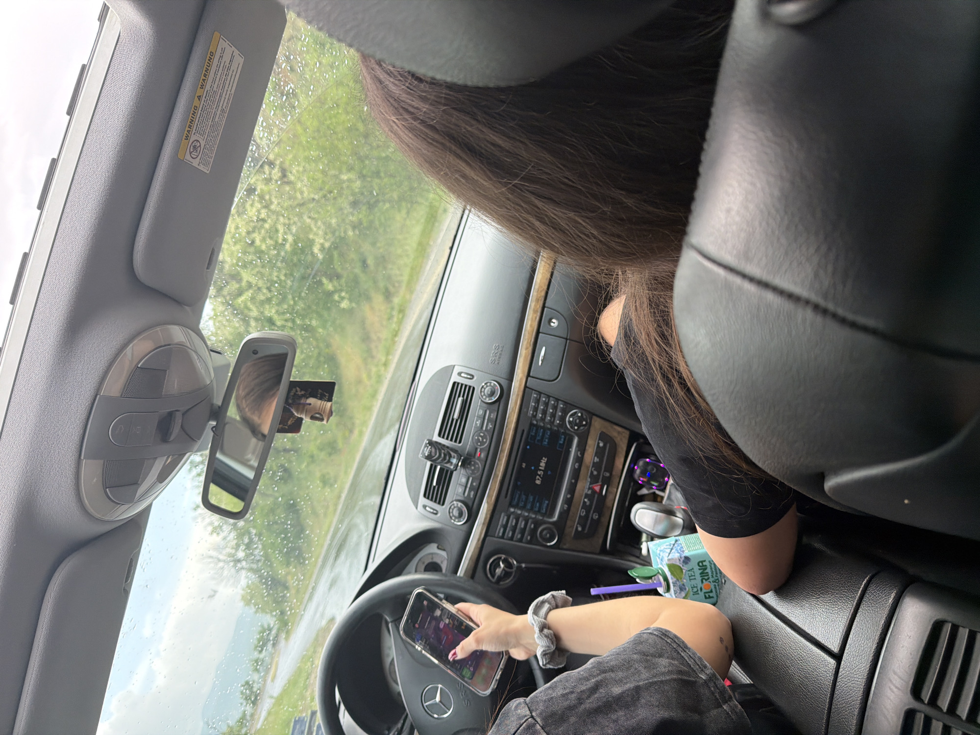 The image depicts the interior of a car, specifically the driver's side. A person is holding a mobile phone in their left hand, resting on the steering wheel. The car's dashboard is visible, displaying the radio and other controls. A drink container is in the center console area. A second person is sitting in the passenger seat. The scene appears to be on a road, visible through the windshield and side windows. The location information provided is Belo Pole, Bulgaria.