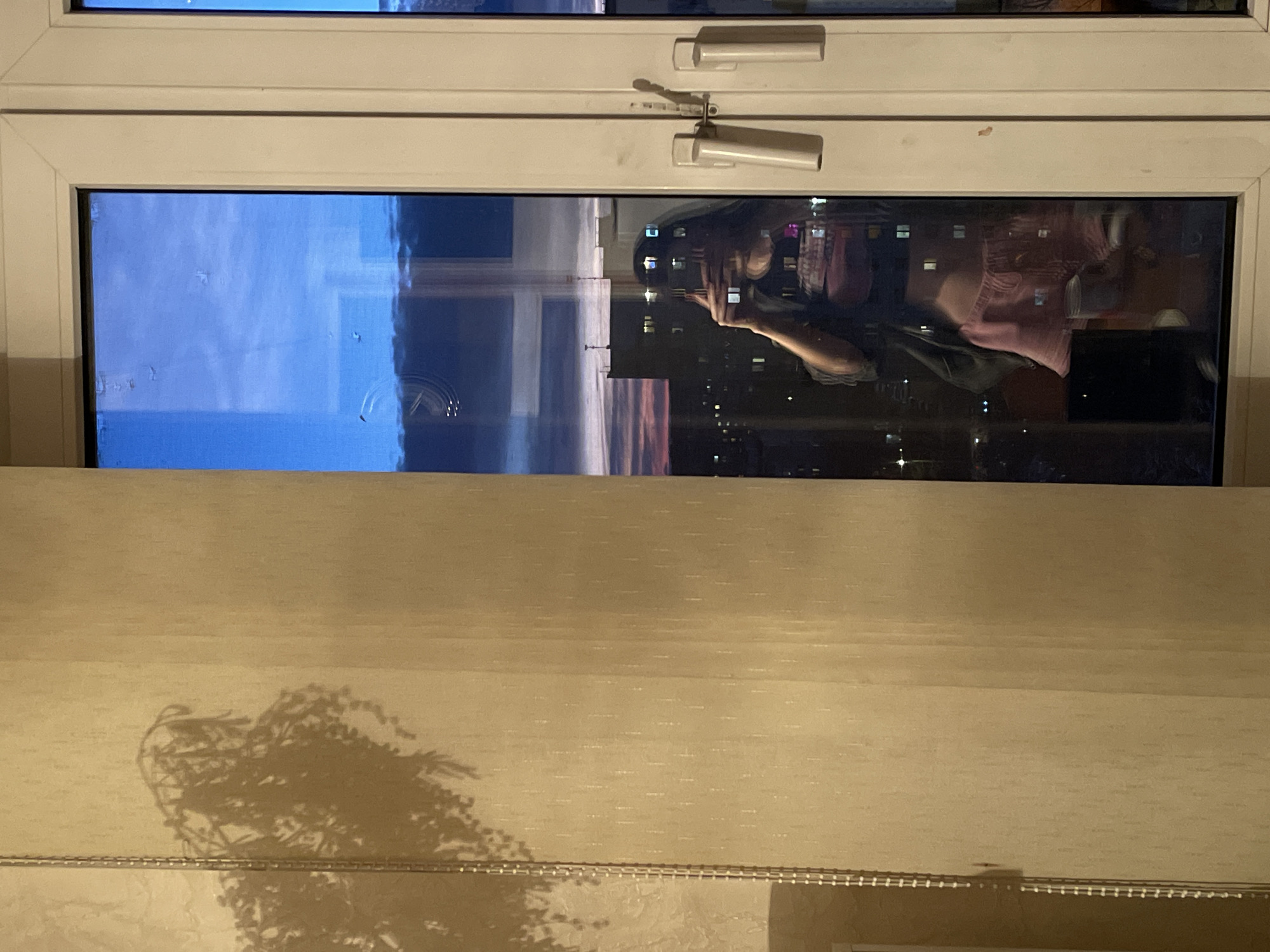 The image depicts a view through a closed white-framed window. Reflected in the glass is a person holding a camera or phone, presumably taking a picture or video. The reflection also shows a cityscape at night, with numerous lights suggesting buildings and infrastructure. Outside the window, a blue sky and the horizon can be seen. Below the window is a drawn shade or curtain, casting a shadow of a leafy plant. Considering the person taking the photo with the phone/camera and looking at the reflection in the window the location seems to be Moscow, Russia.
