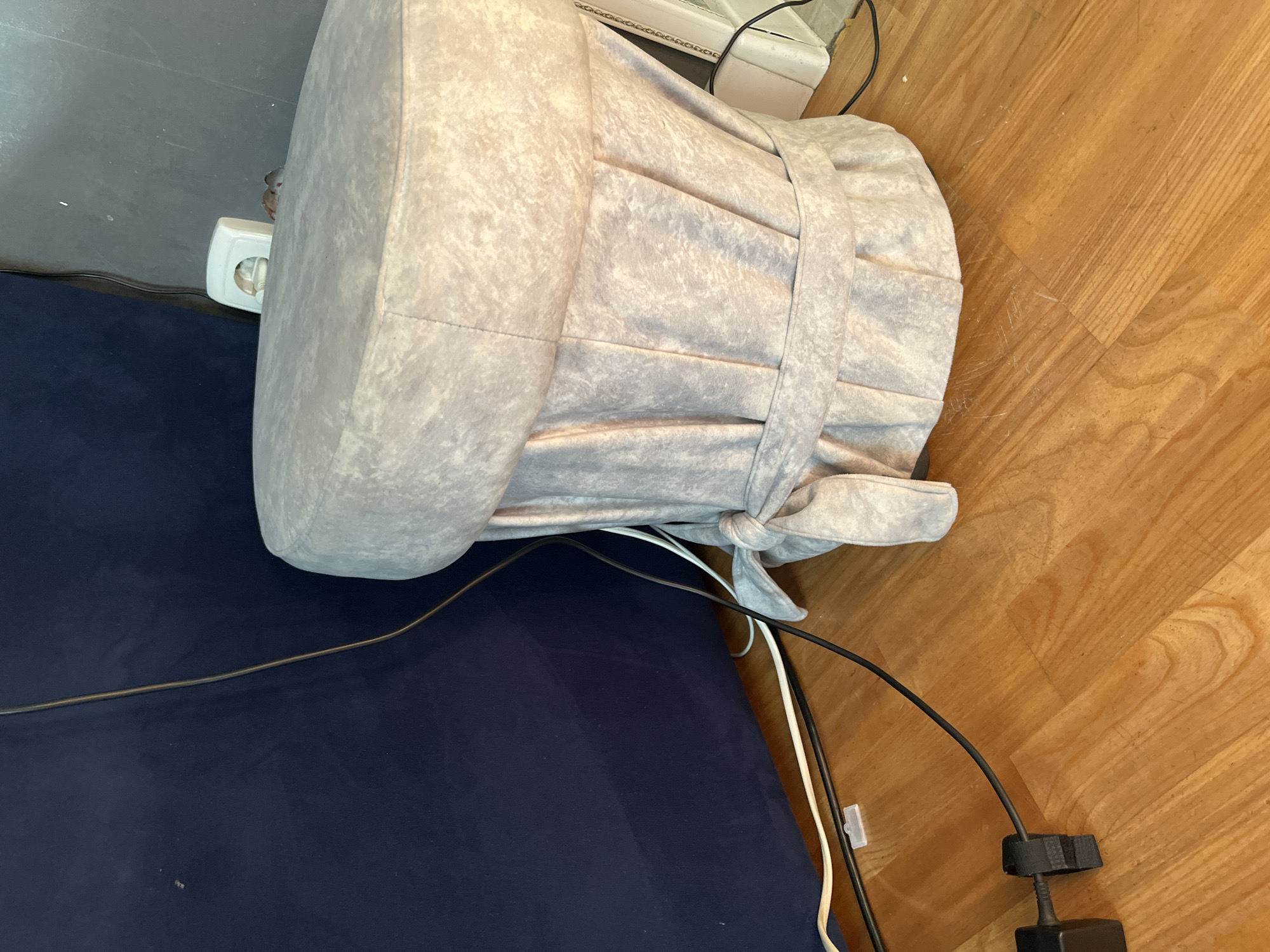 A light gray, pleated ottoman shaped like a chef's hat sits on a wooden floor next to a dark blue upholstered piece of furniture. Black and white electrical cords are visible on the floor. A white electrical outlet is mounted on the wall behind the ottoman. The scene is indoors, with no specific indication of location within Saratov, Russia.