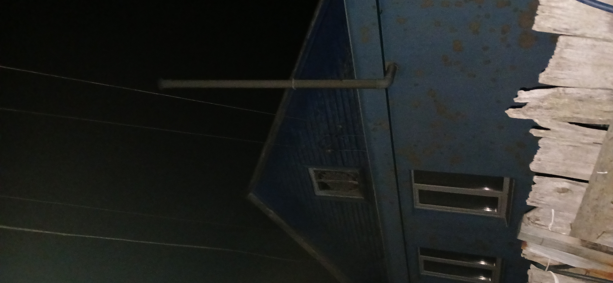 A nighttime exterior shot captures the upper portion of a dark blue, wooden house with a pitched roof and a single dormer window. The house has two prominent windows on the lower level and a smaller window in the dormer. A metal chimney pipe extends vertically from the roofline. Multiple thin, dark lines, possibly electrical wires, stretch across the dark sky above. In the foreground, a rustic, weathered wooden fence runs horizontally across the bottom of the frame. The overall scene suggests a rural or suburban setting in Sumy, Ukraine, likely during nighttime given the dark sky and reflections in the windows. There are no people visible in the image.