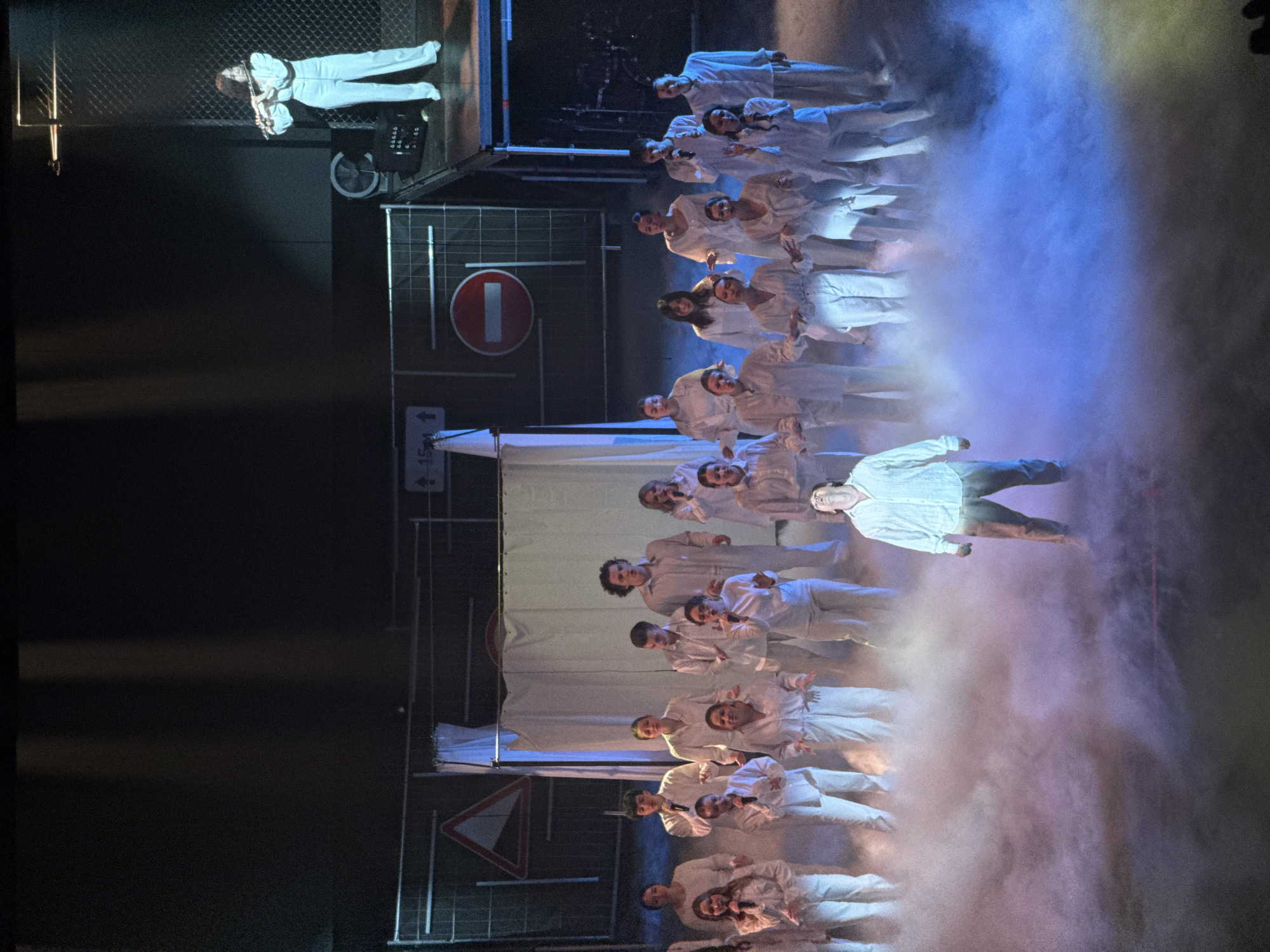 A theatre scene shows performers in white clothing, some singing into microphones. They are on a stage set that includes road signs and fencing. One performer is elevated on a platform. There is smoke or fog effect present. This location is in Perm, Russia.