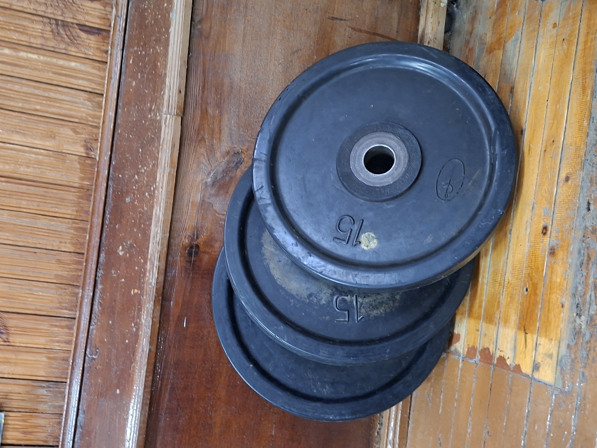 The image shows four black weight plates stacked on a wooden floor. The weight plates have a central hole for a barbell and are marked with "15" indicating their weight in kilograms. The plates appear to be made of iron and are slightly worn. The floor is made of wooden planks with a visible grain and some scuff marks. In the background, there is a wooden wall or structure with vertical slats. The lighting suggests it is daytime, and the scene is indoors. There are no people present in the image. The image does not provide any information that would allow for the determination of its location in the city of Kazan, Russia.