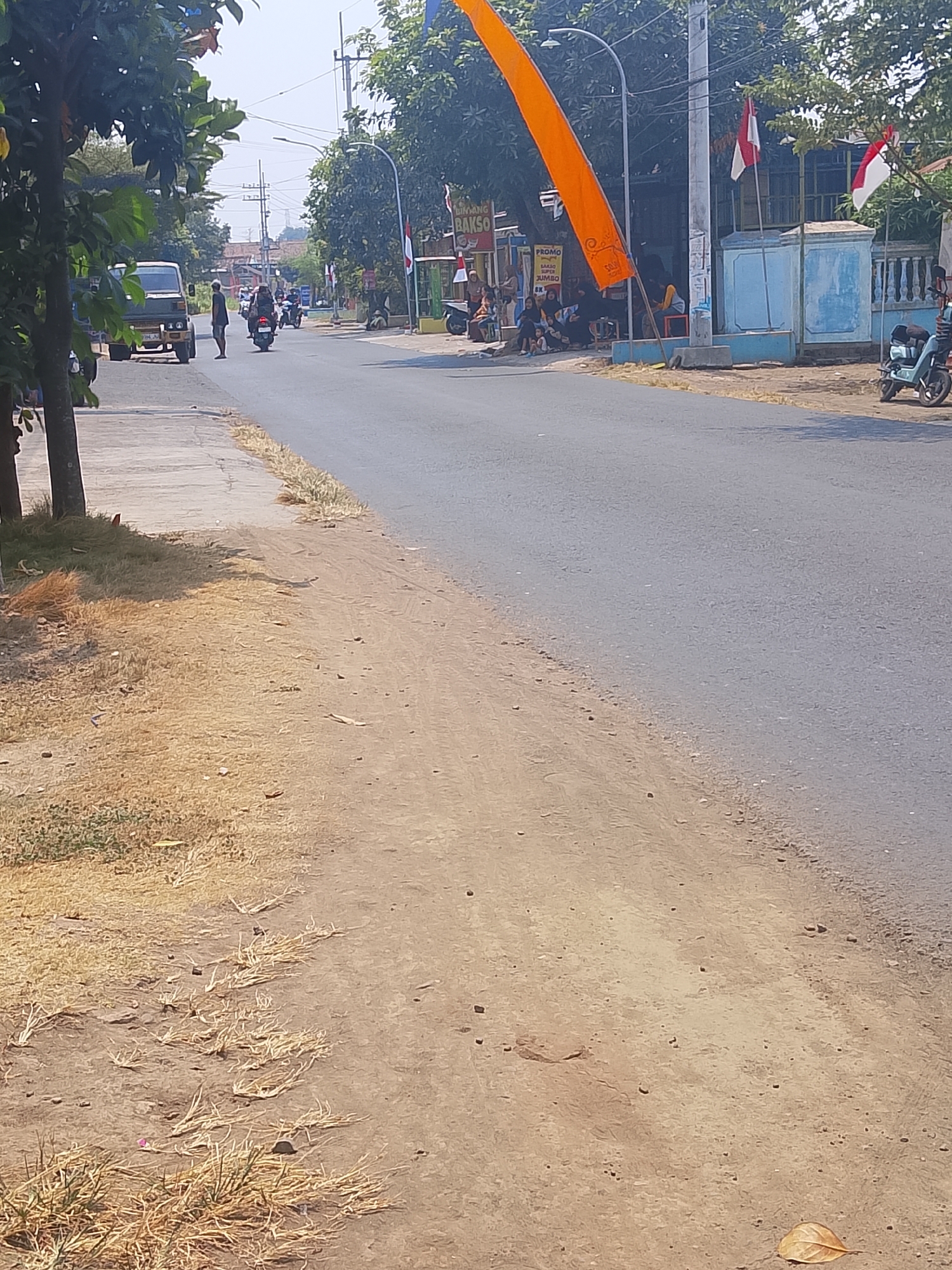 The image shows a street scene in Bangsri, Indonesia, on a sunny day. The road surface is asphalt, with a dry, dusty shoulder on the left side. On the left side of the road, there is a sidewalk and a tree. Further down the road, a small truck is parked. Several people are on motorcycles traveling in both directions on the road. On the right side of the road, there are buildings with storefronts, some with Indonesian flags displayed. An orange flag is prominently displayed on the left side of the road, near a group of people sitting on the ground and on a bench outside a shop. A light blue scooter is parked on the right side of the road. The overall impression is of a typical village street with moderate activity.