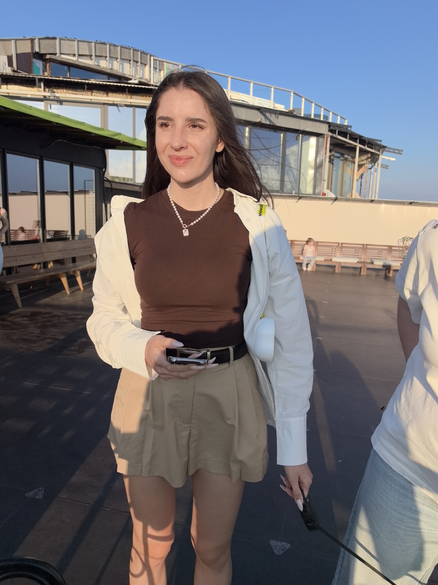 The image shows a young woman standing on an outdoor deck, possibly a rooftop or pier, with a building and a clear blue sky in the background. She is wearing a brown t-shirt, tan shorts, and an unbuttoned white shirt. She has dark hair, a pearl necklace, and is holding a smartphone. The setting appears to be a public area with benches visible in the background. The lighting suggests it is either early morning or late afternoon. There is no discernible information in the image to suggest the location is Tolyatti, Russia.
