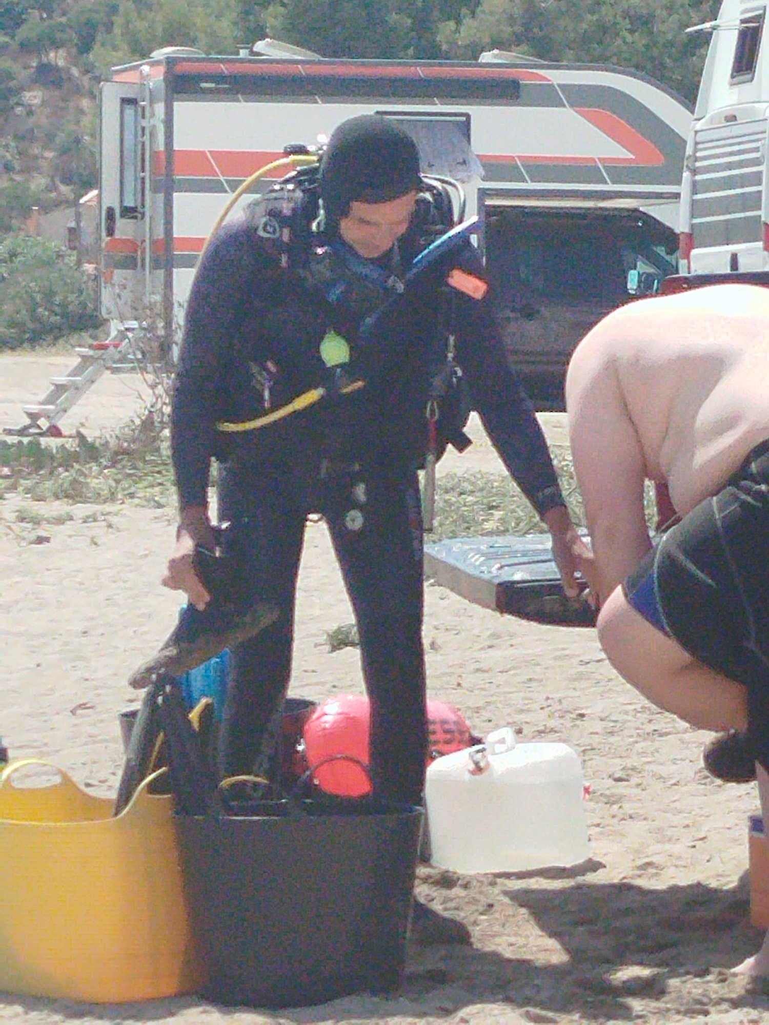 The image shows a diver in full gear on a sandy beach. The diver is wearing a black wetsuit, a black hood, and a black mask. The diver's hands are gloved. In front of the diver is a yellow bucket and a black container. Behind the diver, there is a trailer or camper. Another person, partially visible and shirtless, is to the right of the diver. The background consists of trees and a hillside. The scene appears to be set in a coastal area, possibly during daytime given the lighting.