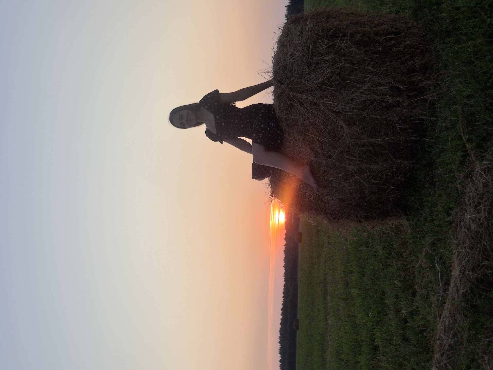 A young woman in a dark dress with a floral pattern sits on a large, round bale of hay in a field at sunset. The sun is low on the horizon, casting a warm orange glow across the sky and illuminating the field with golden light. The woman is silhouetted against the sunset, her face partially obscured by shadow. She is barefoot, with her legs bent and crossed at the ankles. The hay bale is rough and textured, composed of dry straw. In the background, a line of trees forms a dark silhouette against the colorful sky. Other hay bales are visible in the distance, scattered across the green field. The overall scene depicts a serene and picturesque rural landscape during twilight.