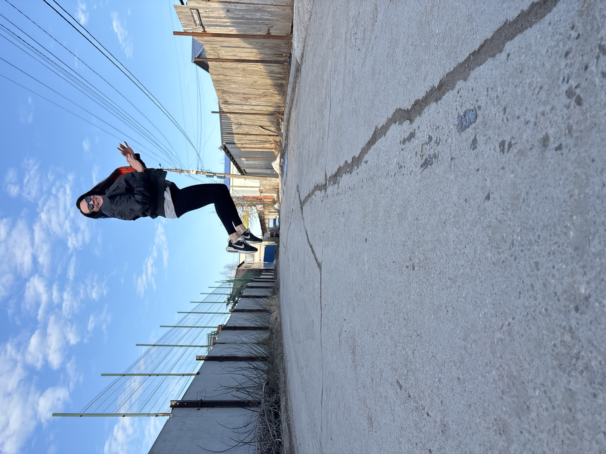 A woman with long dark hair, sunglasses, a dark jacket, and black pants is positioned horizontally in the image, as if suspended in mid-air. Her arms are raised, and she is smiling. The background appears to be an outdoor urban scene, possibly an alley or side street in Chaikovsky, Russia. The camera has captured the scene with a tilt, giving the impression that the person is floating. A concrete walkway dominates the lower portion of the image, with a large crack visible. A metal fence is along one side. A wooden fence or structure is along the other side. In the background, glimpses of other buildings and structures are visible. Overhead are several power lines against a bright blue sky with some clouds.