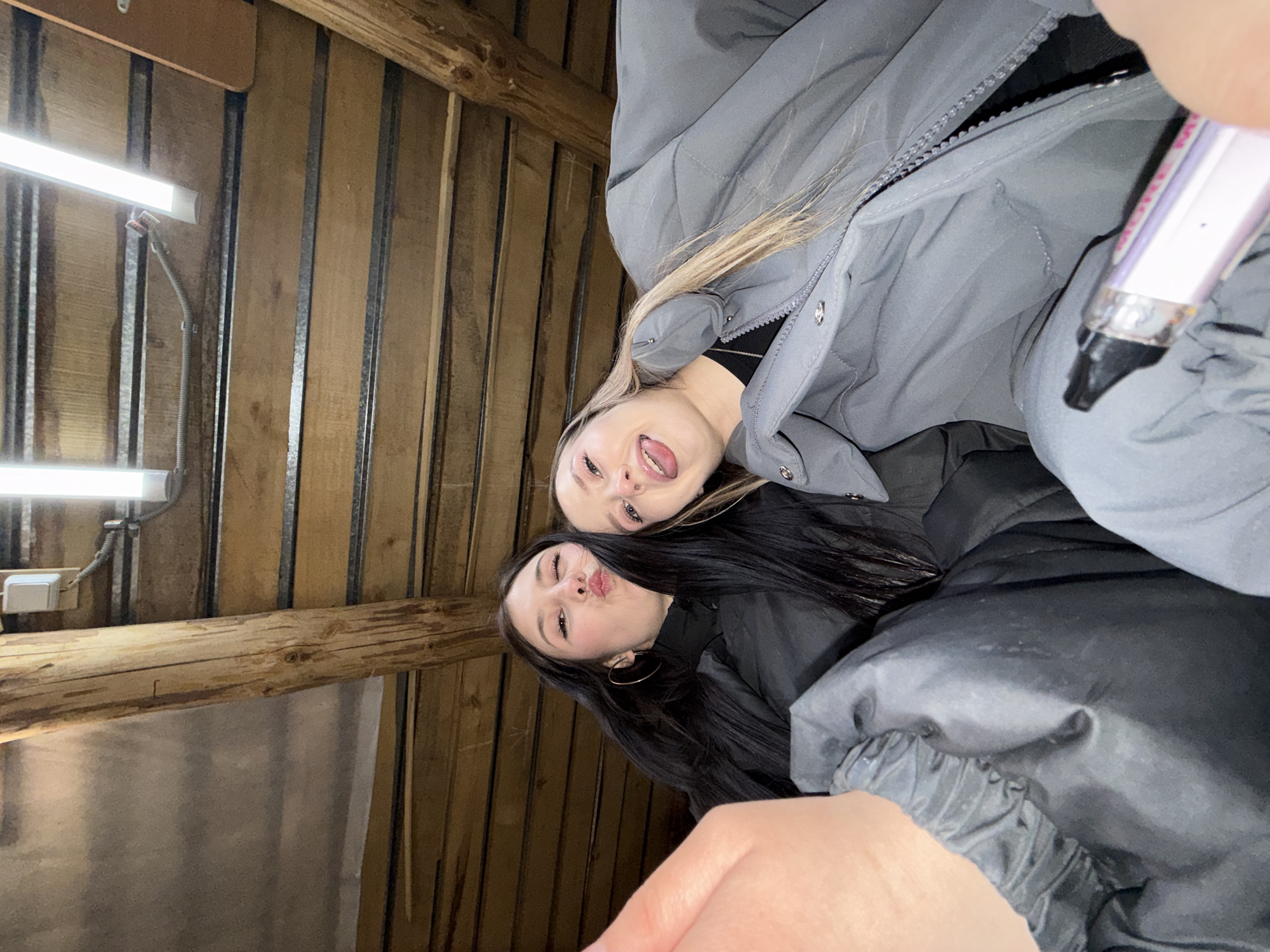 Two women are indoors, likely in a building in Фоки, Russia, based on the location specified. One woman has blonde hair and is wearing a grey puffy jacket, sticking her tongue out. The other woman has long, dark hair and is making a kissing face. Both appear to be posing for the photo. The background features wooden walls and fluorescent lighting. A vape pen is visible in the upper right corner. A hand is visible near the bottom of the image, likely holding the device taking the photo.