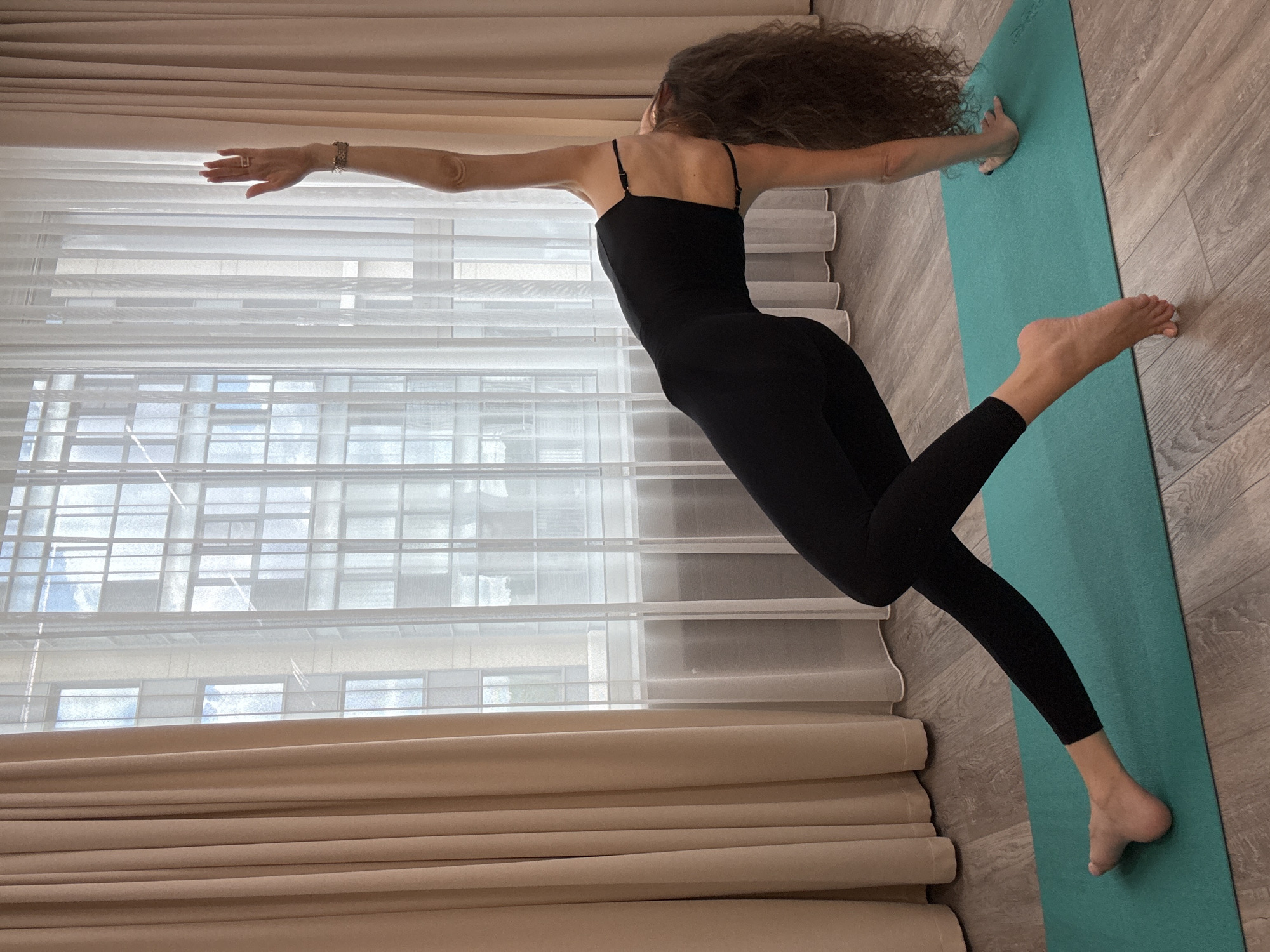 A woman with long, curly brown hair is performing a yoga pose on a teal yoga mat. She is wearing a black full-body leotard and is positioned on her right hand and foot. Her body is arched backward, with her left leg extended and crossed over her right leg. Her right arm is extended straight up towards the ceiling. She is indoors, in what appears to be an apartment, with large windows covered by sheer white curtains and beige drapes. The floor is made of light-colored wood planks. The building visible through the window appears to be a modern residential building, possibly in the city of Khimki, Russia, as indicated by the context.