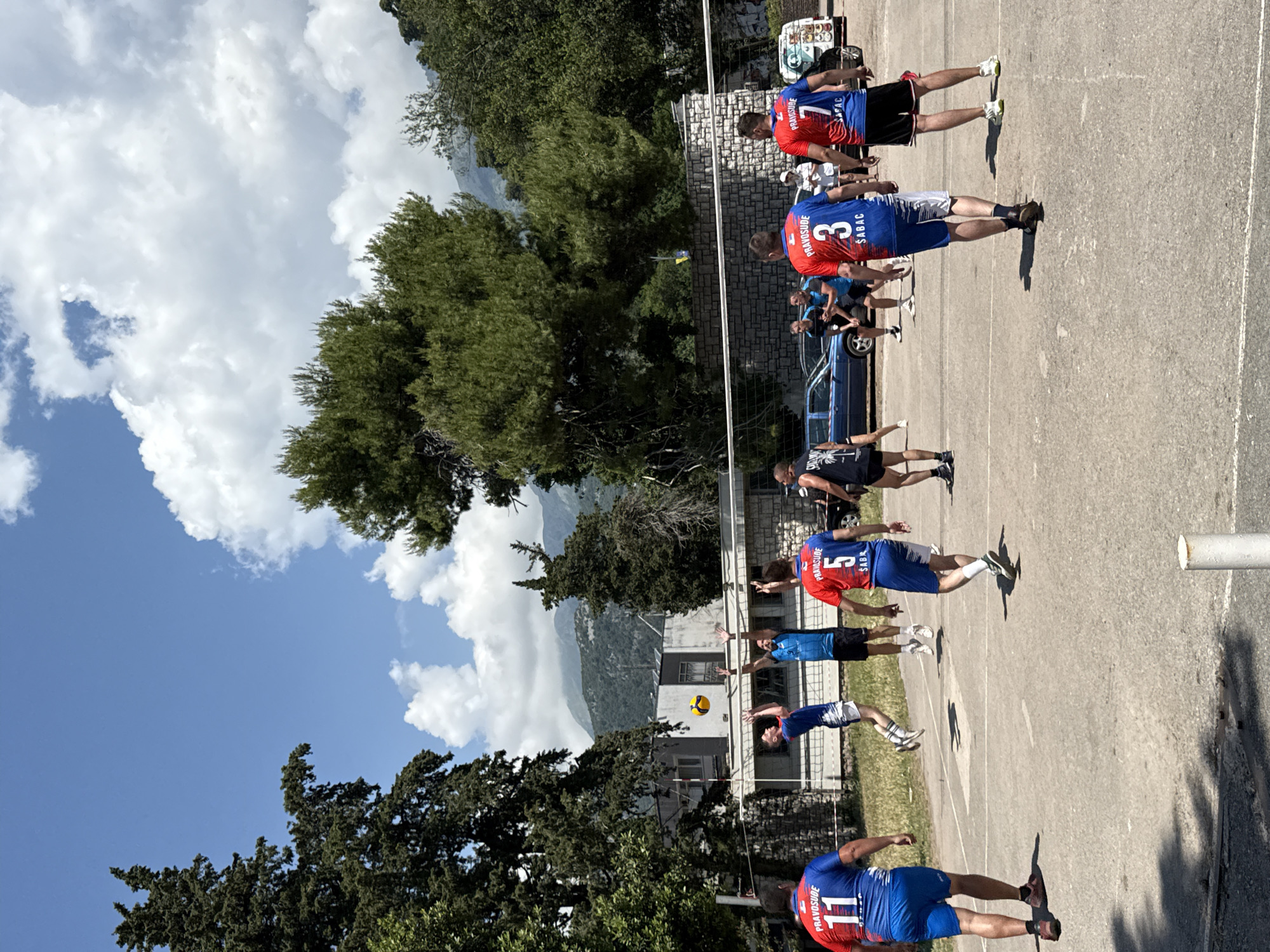 A group of men are playing an outdoor volleyball match on a concrete court in Boreti, Montenegro. The court has white lines marking boundaries. A net is set up across the middle of the court. The players are wearing athletic apparel, with some wearing jerseys with numbers and names. In the background, there are trees, a building, parked cars, and mountains. The sky is blue with some white clouds. The overall scene suggests a recreational sports activity in a scenic, possibly coastal, environment.