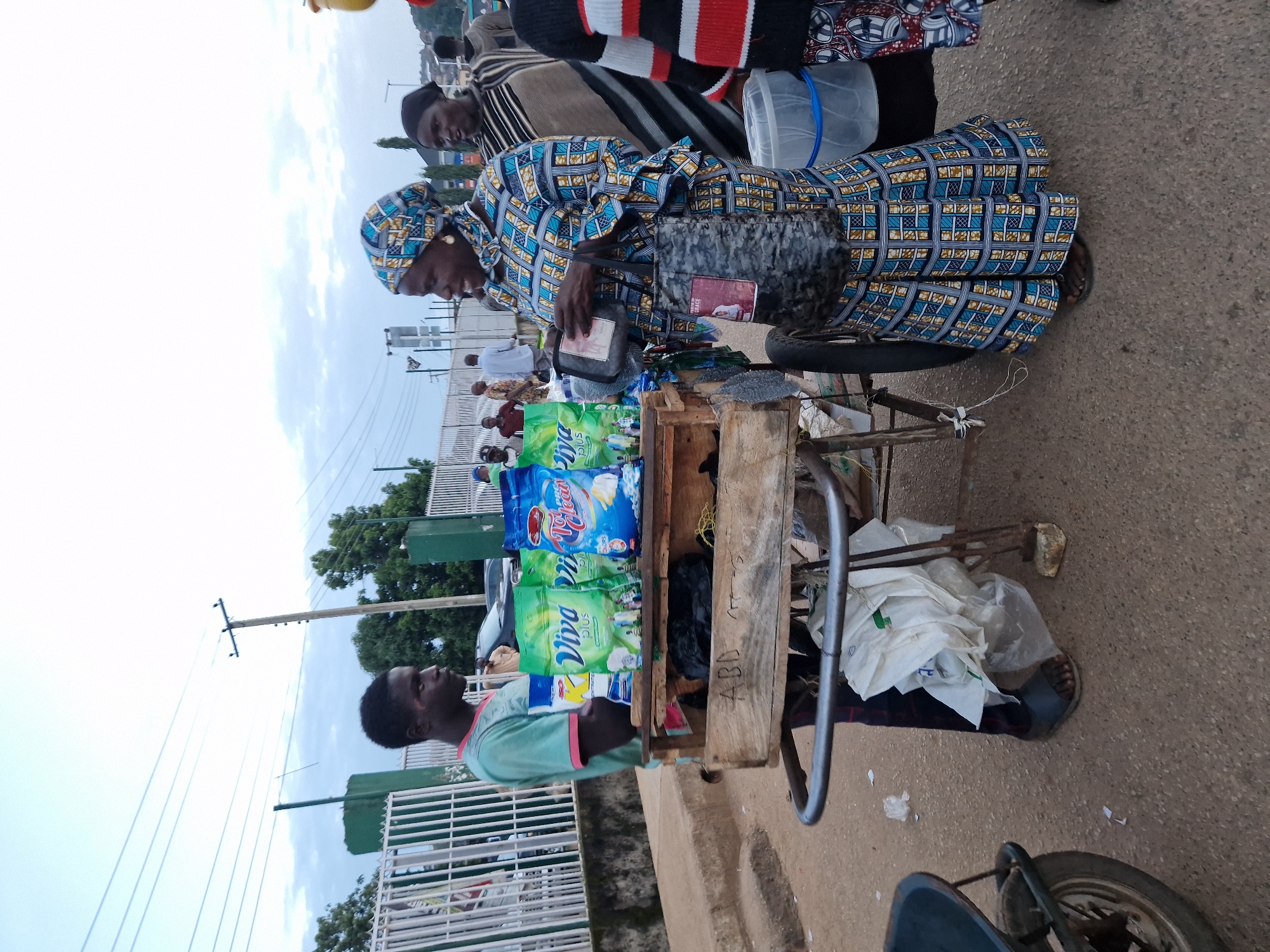 A street vendor, a young man in a teal t-shirt and dark pants, stands behind a wooden cart loaded with bags of laundry detergent at a market stall in Kabong, Nigeria. He appears to be interacting with a female customer, who is wearing a vibrant blue and yellow patterned dress and headscarf. She is holding a small pouch and a larger, patterned bag with a picture on it. In the background, other market-goers and vehicles are visible, and utility poles and wires crisscross the sky. The ground is paved, and a wall with a white fence lines the left side of the image. The overall scene depicts a typical street market transaction in an urban Nigerian setting.