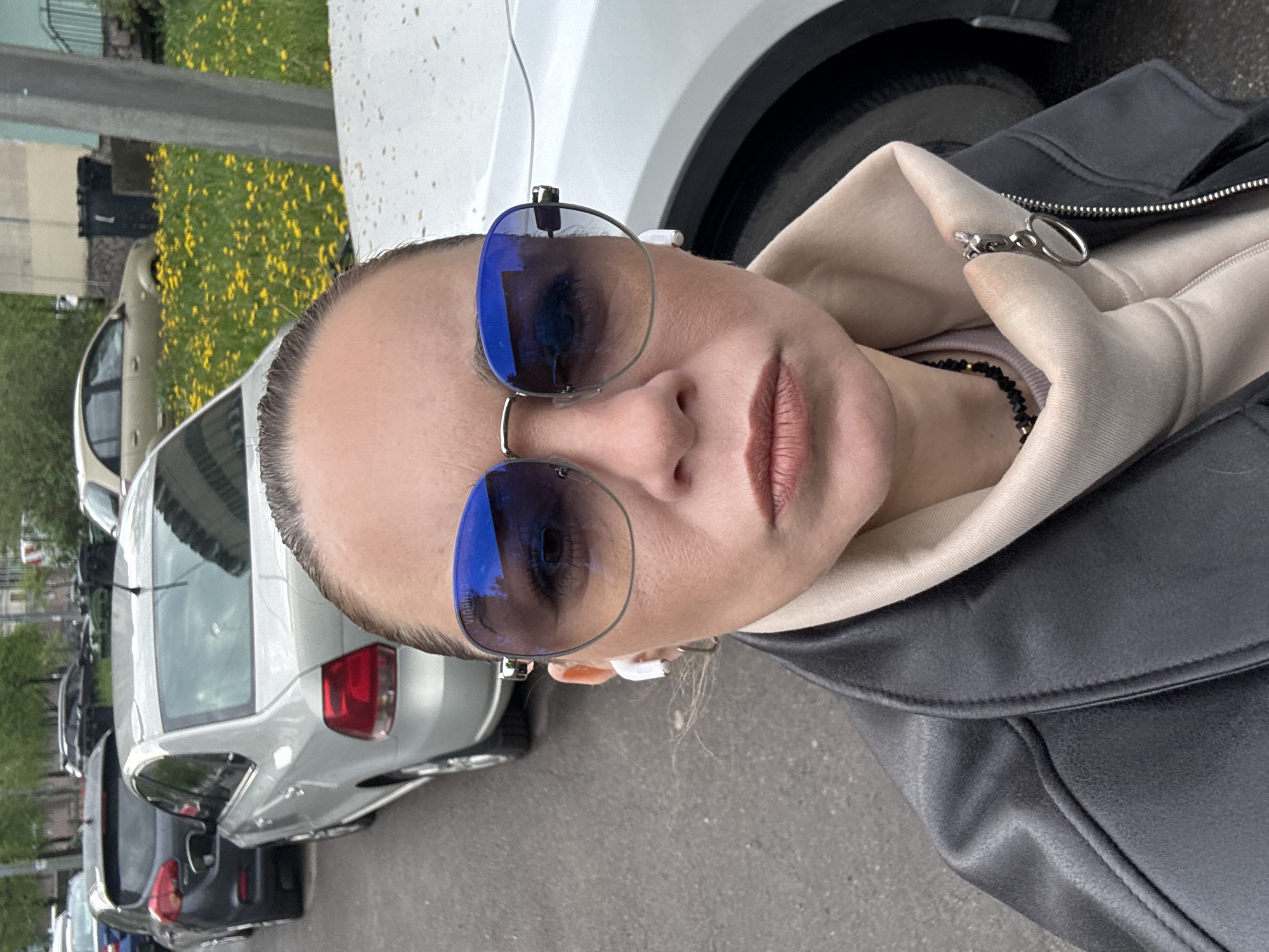 A woman is taking a selfie in an urban setting, likely Saint Petersburg, Russia, given the presence of Russian license plates on several parked cars in the background. She is wearing sunglasses with blue lenses, a beige hoodie, and a black leather jacket. The foreground is dominated by the woman's face, as well as parts of white and gray cars behind her. The environment consists of parked cars along a street, with patches of green grass and yellow flowers visible in the background.