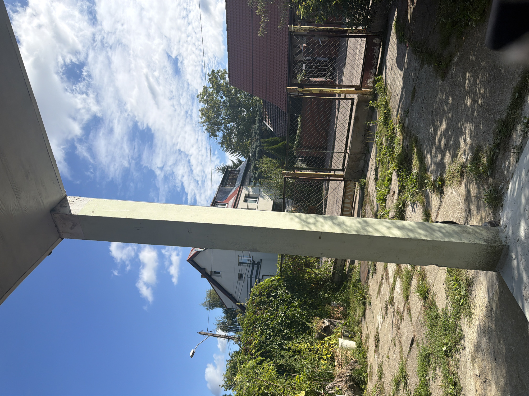 This image depicts a residential area under a bright blue sky with scattered white clouds. The foreground is dominated by a large, light-colored concrete pillar casting a shadow on a paved area with patches of grass. In the midground, a chain-link fence with yellow-painted posts encloses a yard with a red-tiled roof structure behind it. Further back, a multi-story building with white walls and a red-tiled roof is visible, partially obscured by lush green foliage. A street lamp is present to the left of the building. The overall scene suggests a sunny day in a suburban or rural setting, possibly in Szczecin, Poland, given the architectural style of the buildings and the general appearance of the environment. No people or animals are clearly discernible in the image.