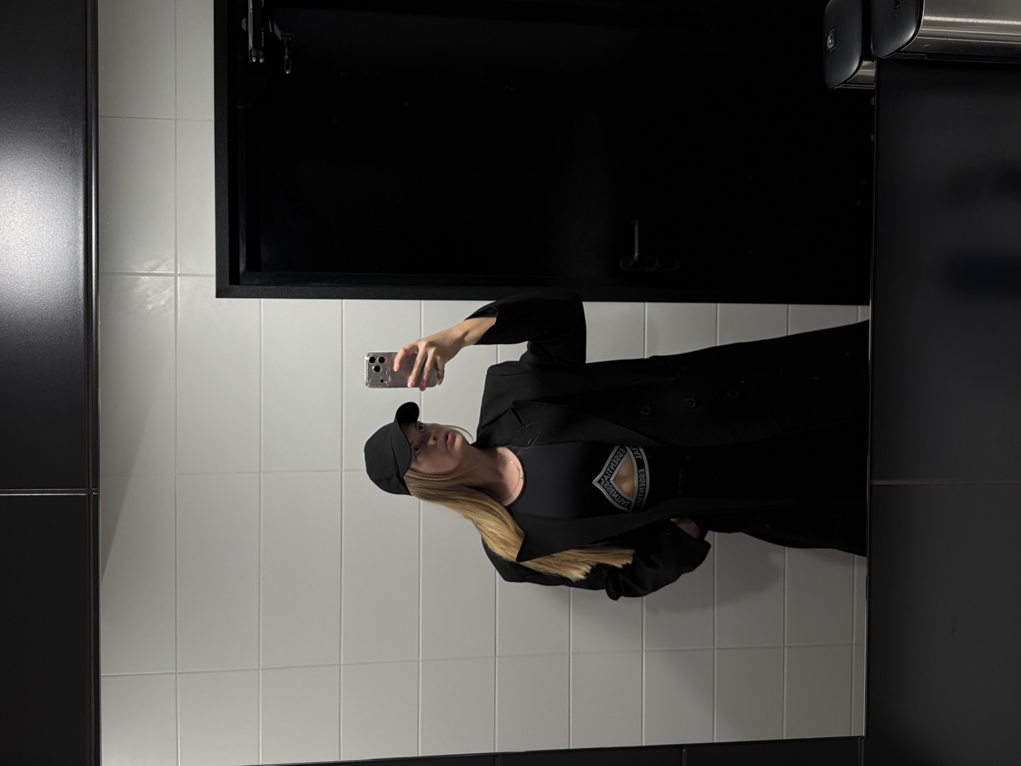 A person wearing a black baseball cap, glasses, a black top, and a black coat is taking a selfie in a bathroom. The bathroom has white tiled walls, a black cabinet, and a stainless-steel dispenser. The location cannot be determined to be Izhevsk, Russia, with only the provided image.