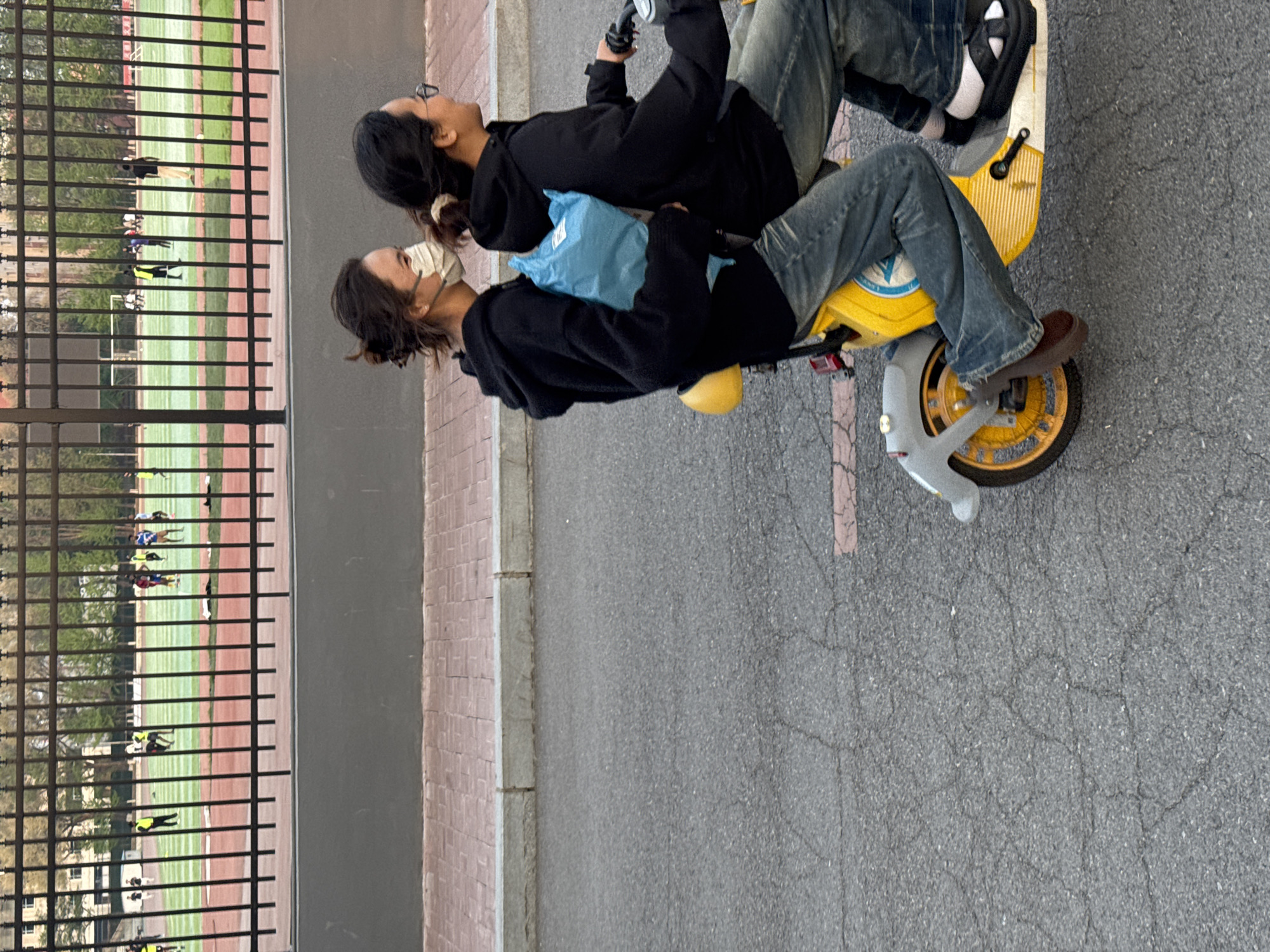 Two individuals on a yellow scooter in Zhongtun, China. The driver, facing the camera, wears a face mask, a black hoodie, and brown shoes. The passenger, behind the driver, wears glasses and a black hoodie. They are riding on a paved road next to a fence that borders a track and field area where people are running.