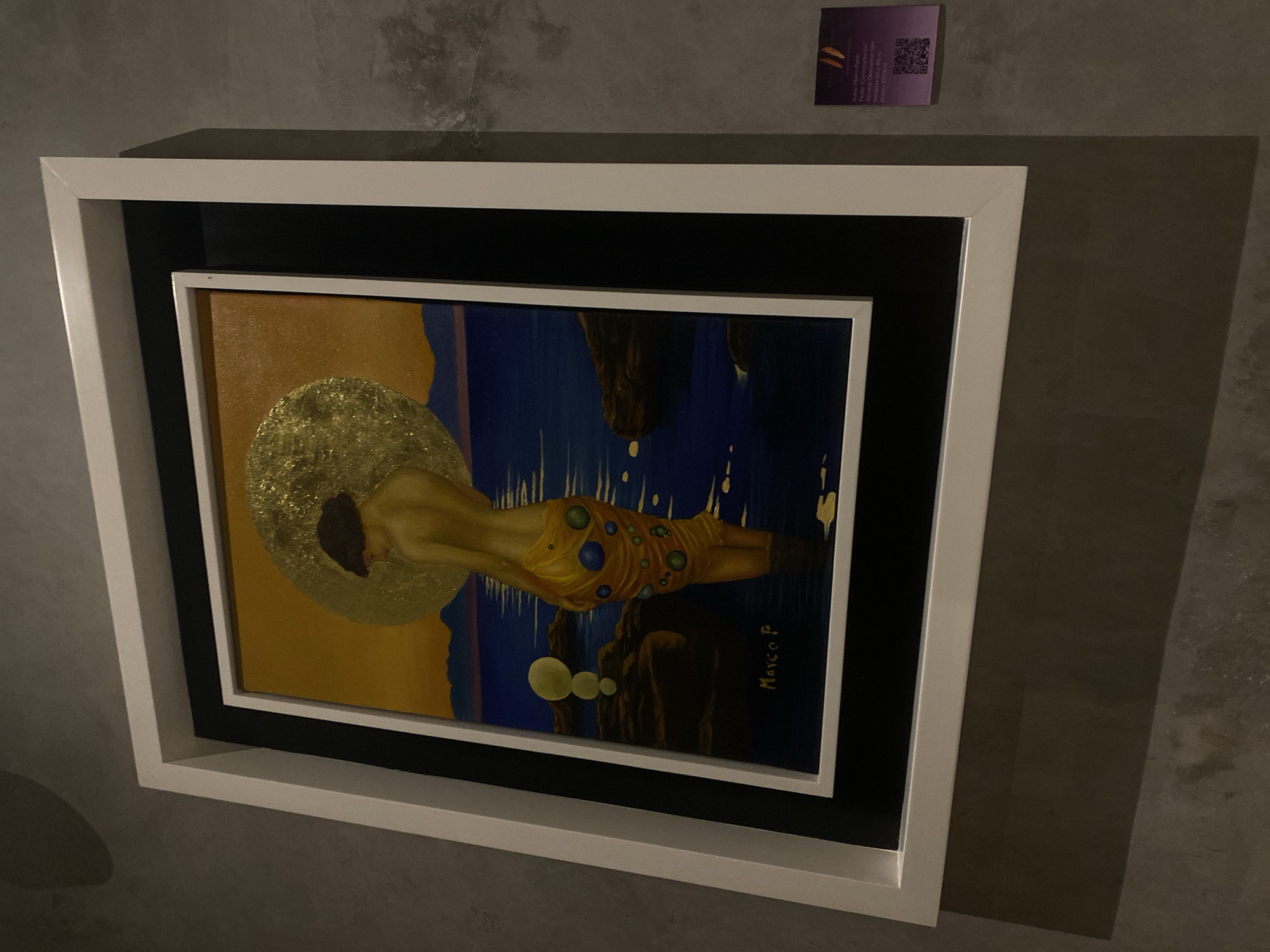 A framed painting hangs on a concrete wall. The painting is in a white frame with a black border. The painting depicts a figure with a golden halo lying on its side. The figure is wearing an orange garment with blue and green circles. The background of the painting is blue with a rocky outcrop. A purple QR code is stuck on the wall above the painting. A shadow is cast on the right side of the wall. The location is in the city of Unknown, Unknown. The label reads "Marco P, Autor Marco Pablo Titulo: "Contemplacion" Tecnica Óleo sobre tela, Medidas 66 x 45cm, Precio $17000.