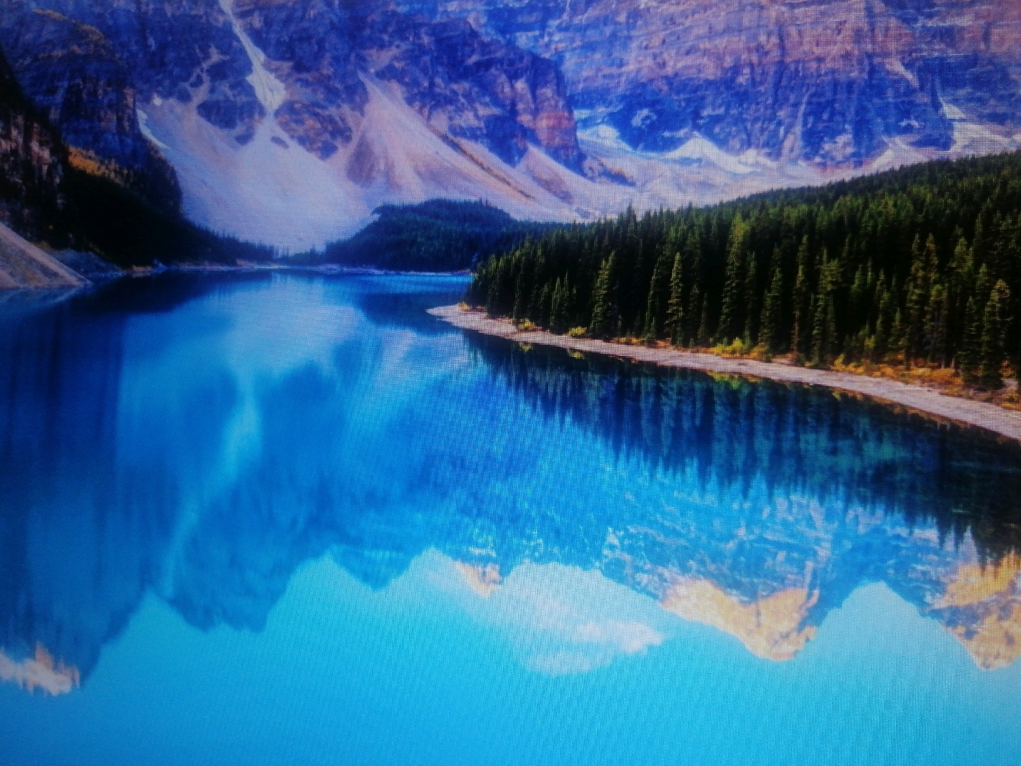 The image shows a breathtaking landscape with a vibrant turquoise lake reflecting majestic mountains and a dense pine forest. The mountains, characterized by their rugged slopes and snow-capped peaks, dominate the background. The forest line hugs the shore of the lake, creating a stark contrast between the dark green of the trees and the bright blue of the water. The reflection of the mountains and trees on the glassy surface of the lake is almost perfect, adding to the serene and picturesque scene. The location is not in the city of Metropolitan Corporation Rawalpindi, Pakistan.